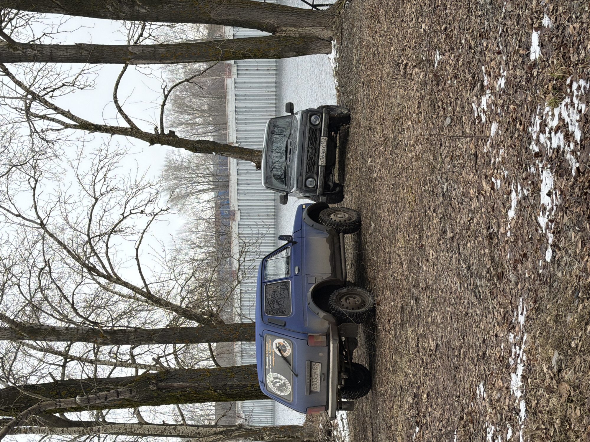 The image shows two vehicles parked on a patch of land covered in fallen leaves and patches of snow. The vehicles appear to be off-road vehicles. A body of water is visible in the background, separated from the vehicles by a barrier. The environment includes bare trees and an overcast sky, typical of a cold season. The location is in the city of Chaikovsky, Russia.