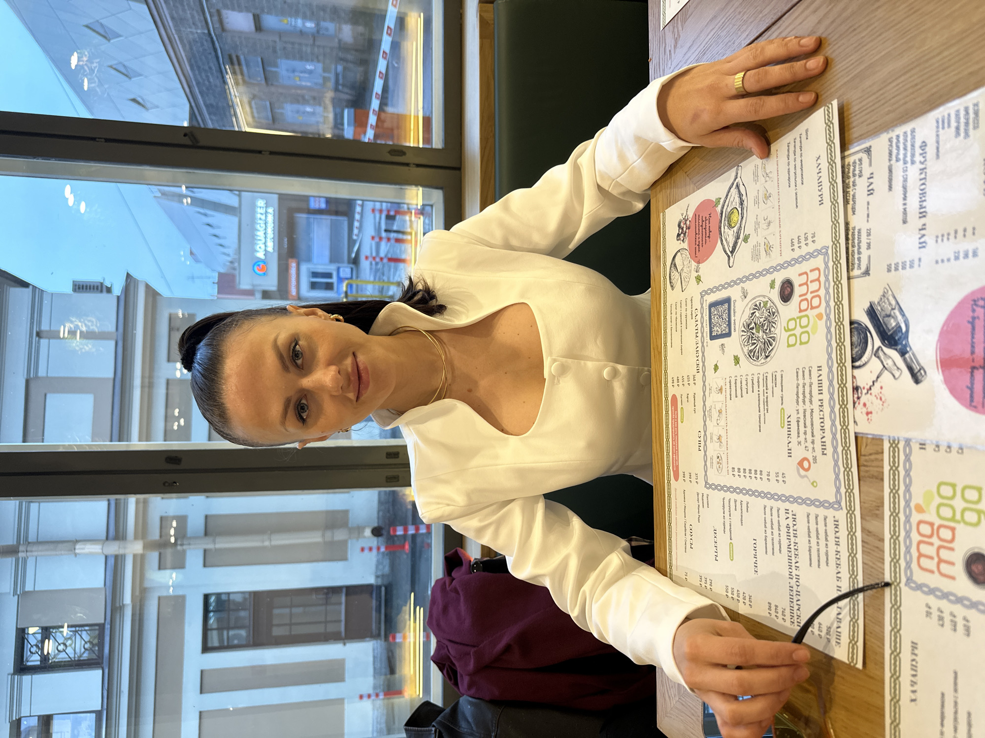 A woman with dark hair pulled back is sitting at a table in a restaurant. She is wearing a white blouse with a plunging neckline and a gold necklace. She is looking at the camera and smiling slightly. Her hands are on the table in front of her, holding a pair of glasses. There are menus on the table, and in the reflection of the window there is a building with a sign that says "AQUAGIZER АВТОМОЙКА" in Russian, which translates to "Car Wash." The restaurant is likely located in Saint Petersburg, Russia, based on the text in the menu and the sign in the window.