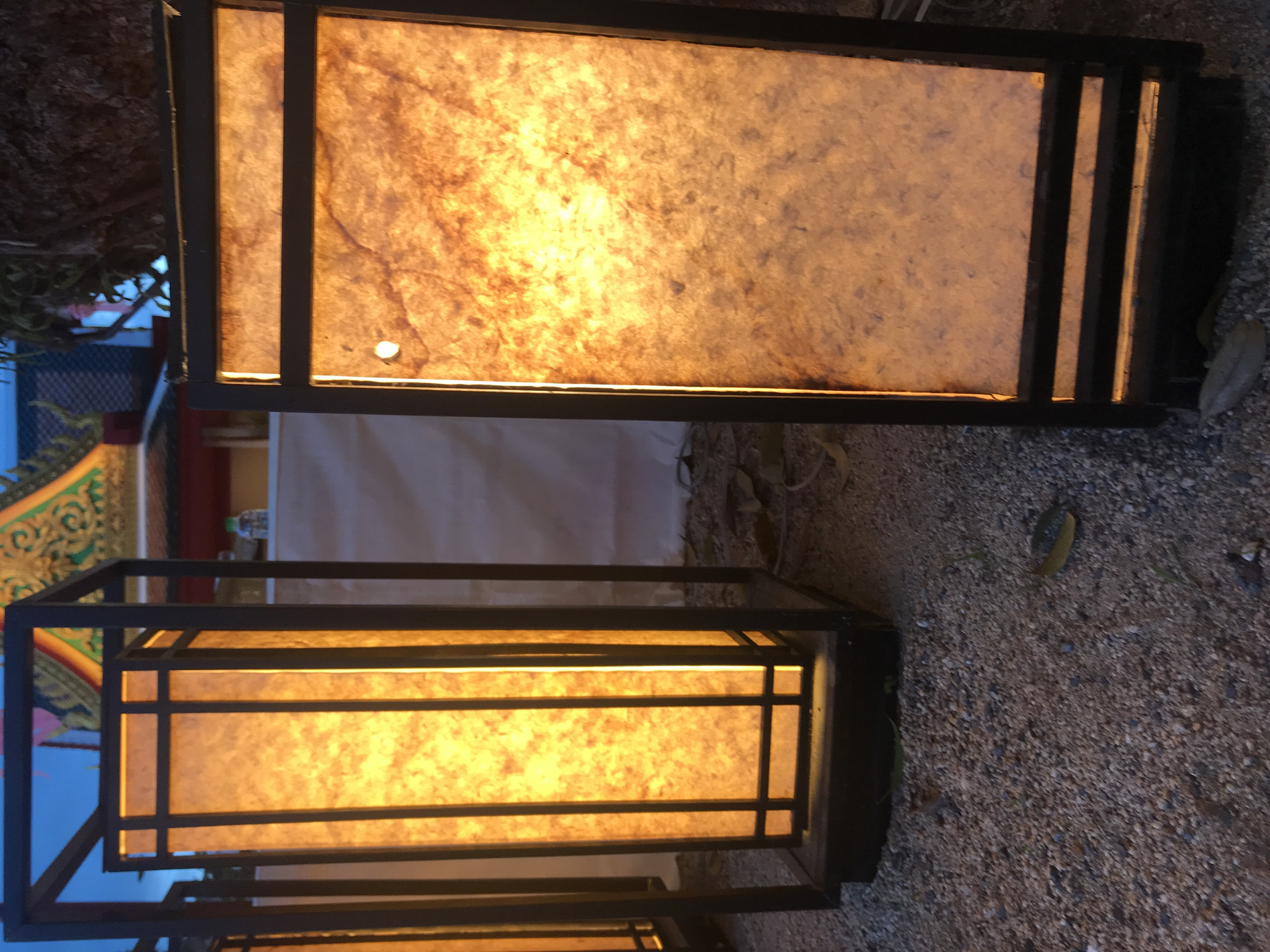 Two lanterns with illuminated yellow-orange paper panels are arranged on a sandy, gravelly surface. The lanterns have a dark, rectangular frame. The upper lantern is oriented horizontally, while the lower lantern is vertical. A building with a green and gold ornate roof is partially visible in the background, with a white tent-like structure behind it. The setting is Baan Mae Nam, Thailand.