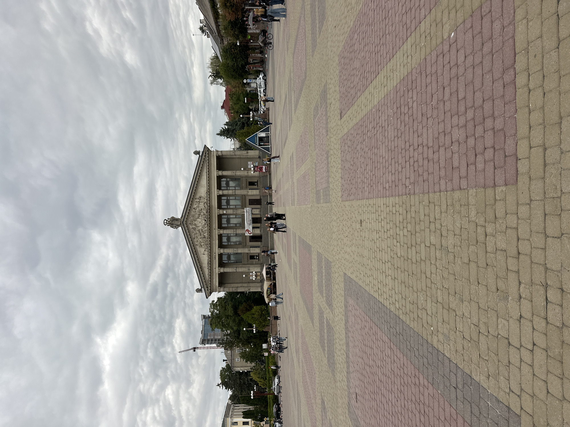 The image shows a wide, open pedestrian square in Ternopil, Ukraine, dominated by a large, classical-style building with columns and a pediment at the center of the frame. The square is paved with interlocking bricks in a pattern of reddish-brown and light beige. Several people are visible walking across the square or standing in small groups. To the left of the central building, there is a multi-story building with windows and a tall construction crane in the background. To the right, there are trees and smaller buildings. The sky is overcast with grey clouds. In the foreground, the patterned brickwork of the square is clearly visible.
