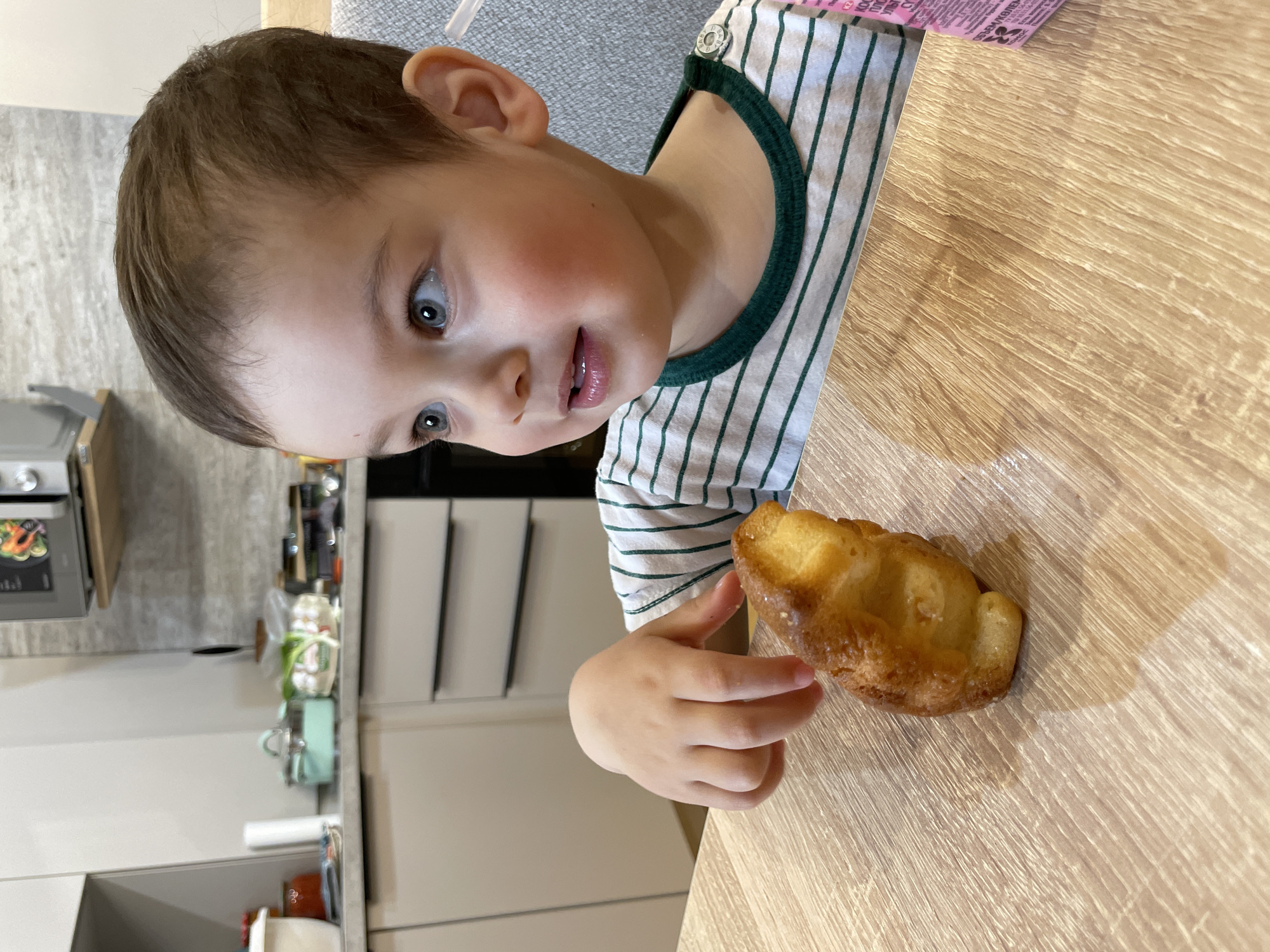 A young child with fair skin, brown hair, and blue eyes is seated at a light brown wooden table. He is wearing a striped shirt with green trim around the neck. He is looking at and pointing towards a golden-brown pastry on the table. Part of a modern kitchen is visible in the background, including gray cabinets, a stainless steel microwave, and various kitchen items. A container of juice is visible behind the child. The image shows a domestic setting, likely a home kitchen. There is no information available to identify the location as Zav'yalovo, Russia.