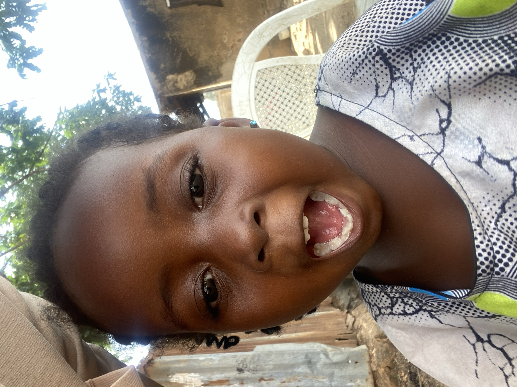 A close-up, rotated portrait captures a young child, likely a girl, from Kuru Dungus, Nigeria. She has dark skin and dark, curly hair, and looks directly into the camera with an animated, open-mouthed expression, revealing her teeth. Her wide, bright eyes convey a sense of joy and playfulness, suggesting she is laughing or shouting playfully. She wears a light-colored garment with a distinctive black and white pattern featuring both geometric dots and crack-like lines, accented with hints of lime green and blue.
The background, mostly out of focus, indicates an outdoor setting during the daytime, with bright sunlight filtering through green tree foliage. Visible behind the child are elements of a rustic environment, including a white plastic chair, parts of a building structure, and what appears to be corrugated metal. The overall impression is one of candidness and cheerfulness in a natural, informal setting. No visible text is legible in the image.