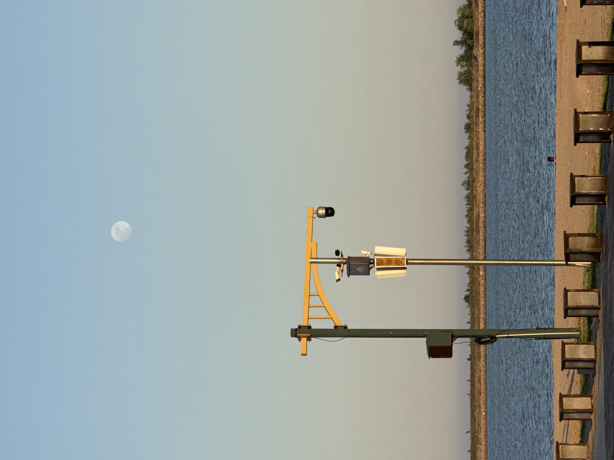 The image depicts a sideways view of a landscape. In the upper right quadrant, there is a body of water with a shoreline and trees. Adjacent to the water are several rectangular structures, potentially benches.
In the middle of the image, a metal structure with various components is visible. This structure consists of poles, beams, and equipment, including a camera and other electronic devices.
In the upper left quadrant, there is a moon against a clear sky. The absence of people, actions, or interactions indicates a static scene. The image does not contain any details that would help to determine the location in Taihe District, China.