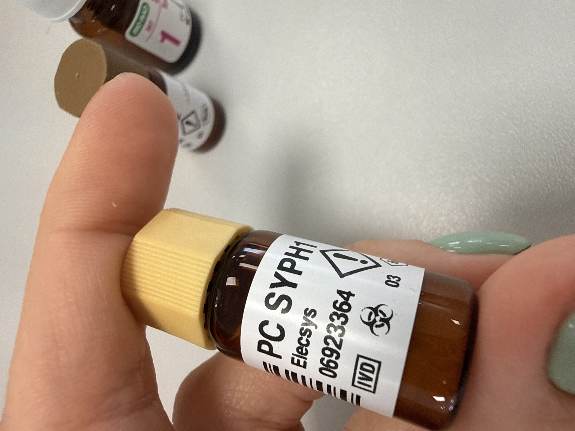 A close-up shot features a human hand holding a small, amber-colored glass vial with a light yellow, ribbed screw cap. The hand, with fingernails painted light green, grips the vial between the thumb and index finger. A white label wraps around the vial, displaying black text and symbols. Prominently visible text includes "PC SYPH1", "Elecsys", a numerical code "06923364", "IVD", and "03". Two warning symbols are also present: a black exclamation mark within a yellow triangle, and a black biohazard symbol.
In the blurred background, several other similar small vials are partially visible on a light-colored, possibly white, surface. One background vial has a white cap and a label with a pink number "1", while another shows a brown cap and a small black warning symbol. The scene is brightly and evenly lit, suggesting an indoor setting like a laboratory or medical facility where such diagnostic reagents or controls are used. The image provides no specific visual cues to identify a geographic location, but it was captured in Moscow, Russia.