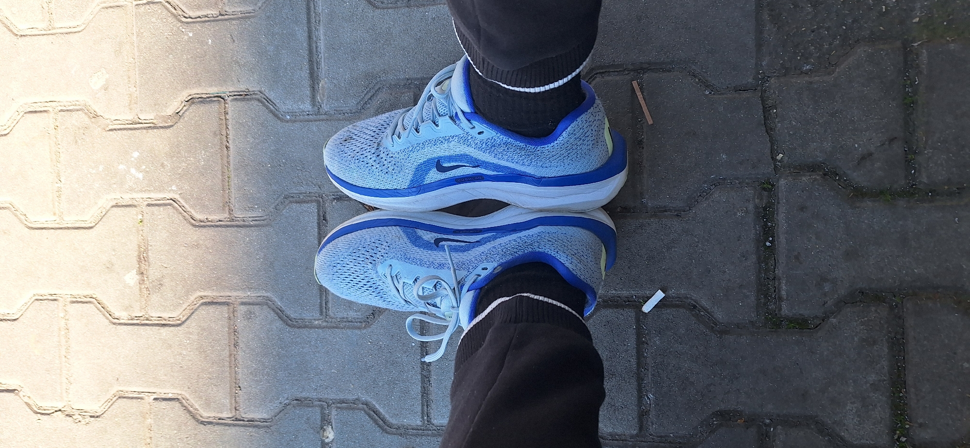 The image shows a pair of blue and white Nike running shoes worn by a person standing on a paved sidewalk. The person is wearing black athletic pants with white stripes on the socks. The sidewalk is made of rectangular interlocking pavers, with some sections appearing lighter than others due to sunlight. There are small pieces of debris visible on the sidewalk around the shoes, including a small piece of wood and a possible cigarette butt. The location is Stara Zagora, Bulgaria, as inferred from the pattern of pavers which are commonly used in this city.