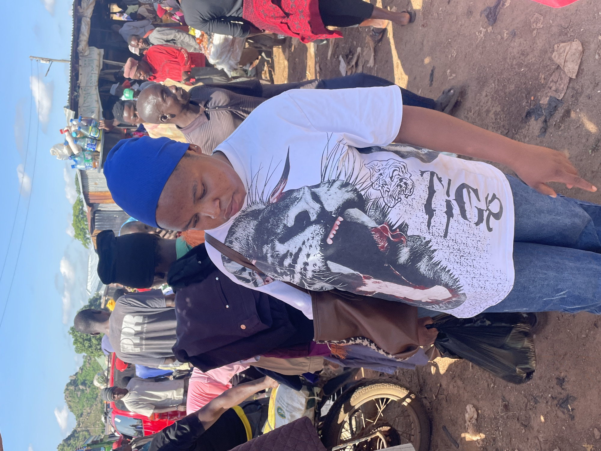 The image depicts a bustling outdoor market scene in Gura Top, Nigeria. A woman wearing a blue head covering and a white t-shirt with a graphic print of a tiger is the central subject, holding a brown bag and a black plastic bag. She is surrounded by other individuals and stalls selling various goods. In the background, there are more people, motorcycles, and makeshift structures, indicative of a lively marketplace. The ground is dusty and unpaved. The overall impression is one of a vibrant and busy commercial hub.