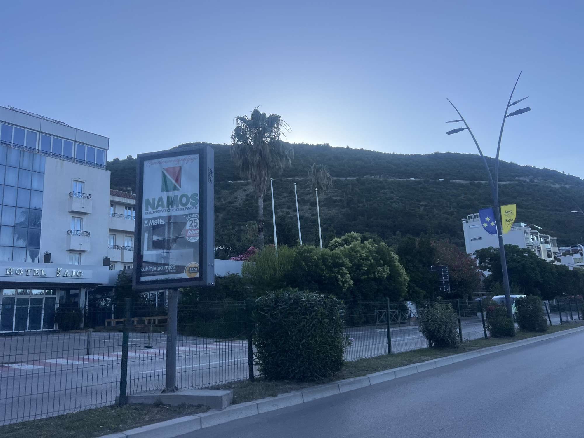 This is a daytime exterior, eye-level, wide shot of a street scene in Budva, Montenegro. A large billboard for "NAMOS DAJKOVIC COMPANY" stands prominently in the foreground, with a hotel named "HOTEL SAJO" to its left. The hotel is a multi-story building with a modern facade featuring large windows. Behind the billboard and hotel, a lush green hillside rises, with a few scattered palm trees and dense vegetation. The sky is a clear, pale blue, with the sun visible near the horizon, creating a soft glow. The street itself is paved and appears to be relatively empty, with a metal fence lining the sidewalk. In the distance, more buildings can be seen, including a white building with blue and yellow flags, suggesting a commercial or public area. The overall impression is one of a developing urban environment nestled against a natural landscape.