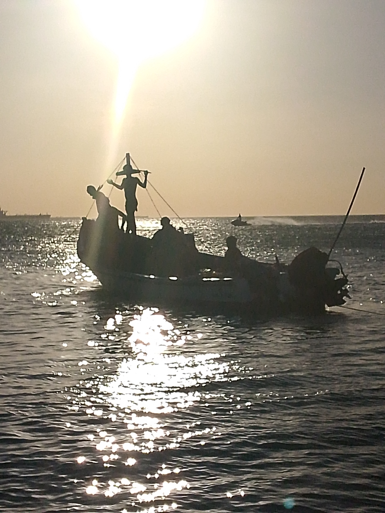 A low-angle shot of a small boat with several silhouetted figures on board, taken in what appears to be Villa Marina, Venezuela. The bright sun is high in the sky, creating a silhouette of the boat and its occupants. In the foreground, the water reflects the sunlight. A figure stands atop the boat, silhouetted against the sun. There are other figures seated and standing inside the boat. The boat has a mast-like structure at the bow and an outboard motor at the stern. In the background, other vessels are visible on the water, one of which is small enough to be a jet ski.