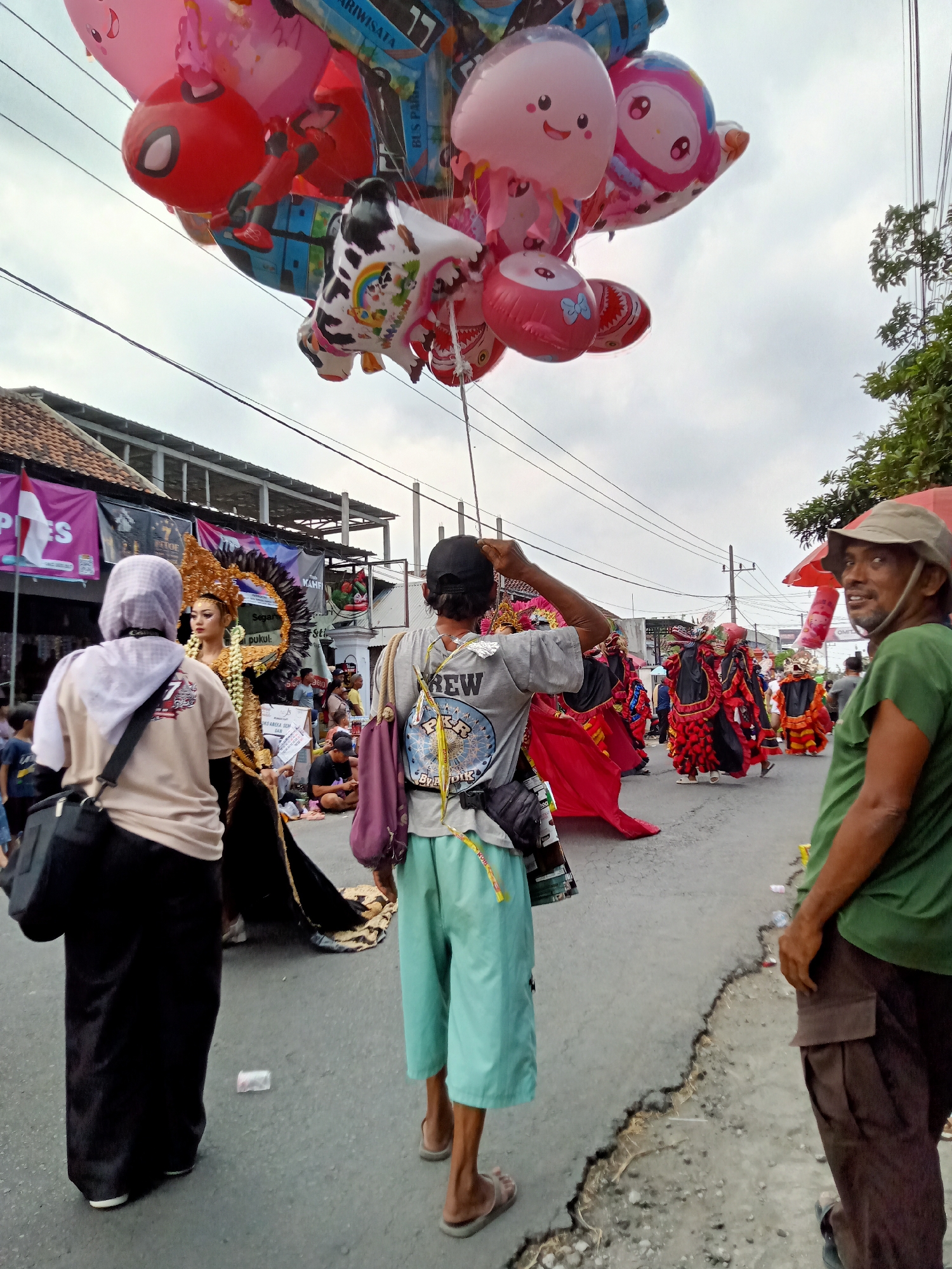 On July 18, 2023, a street parade is taking place in Simo, Indonesia. A large cluster of balloons, including a Spiderman-themed balloon, is held by an unseen person. In the foreground, a woman in a beige shirt and black skirt stands facing a performer in an elaborate costume adorned with gold and feathers. To the right, a man wearing a green shirt and a hat is looking towards the camera. Behind the main subjects, other participants in colorful costumes are visible, as are spectators and buildings lining the street. The atmosphere suggests a festive or cultural event.