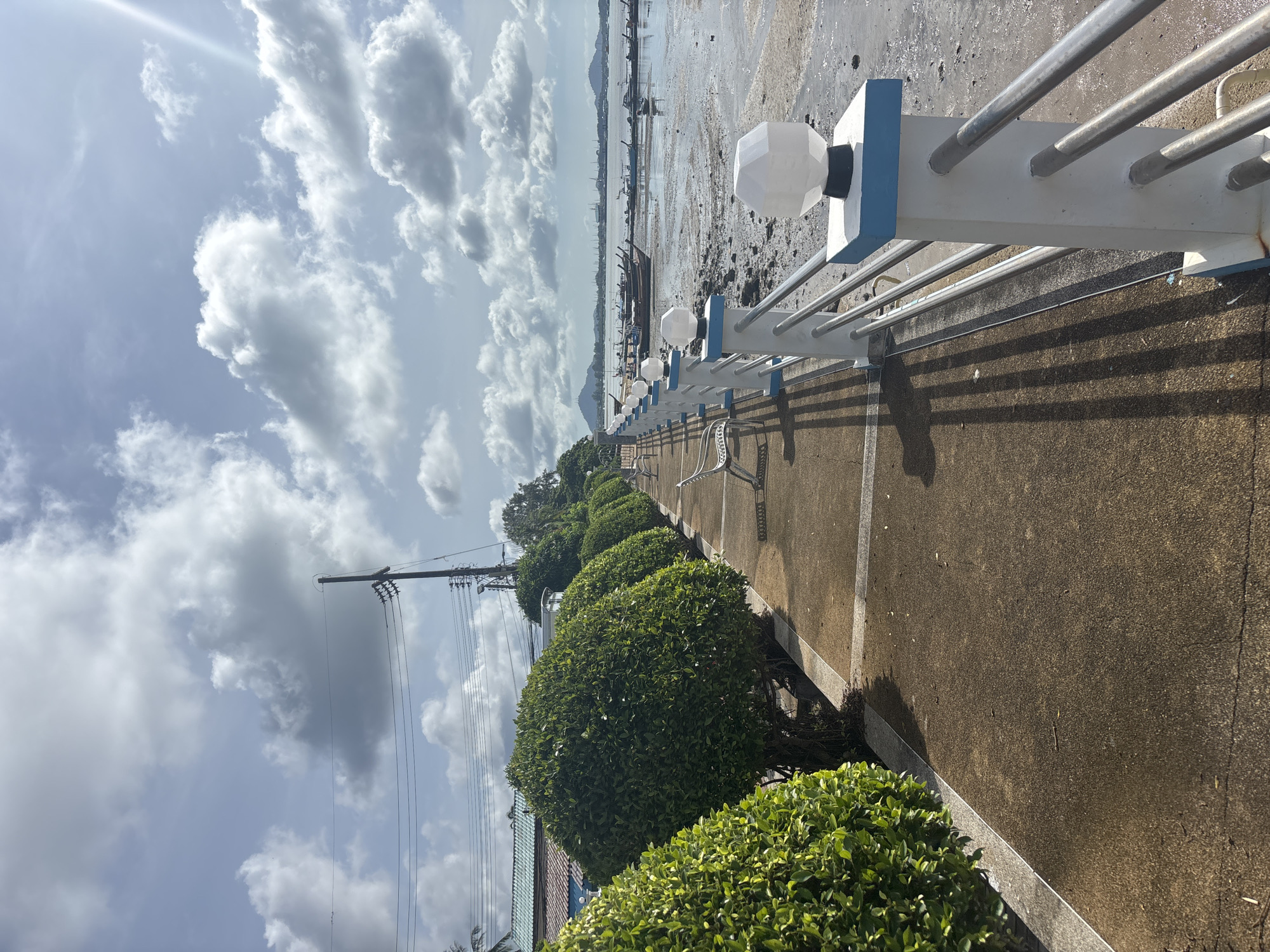 The image depicts a paved walkway along a waterfront in Rawai, Thailand. The walkway is lined on the left by a row of manicured, spherical green bushes. On the right, a white railing with blue accents and hexagonal light fixtures separates the walkway from the water. In the distance, numerous longtail boats are moored on the shallow, muddy water, with a hazy mountainous coastline visible beyond. The sky is partly cloudy with sunlight filtering through. A single white bench is visible on the walkway. Telephone poles and wires are present in the background, near some buildings with blue roofs.