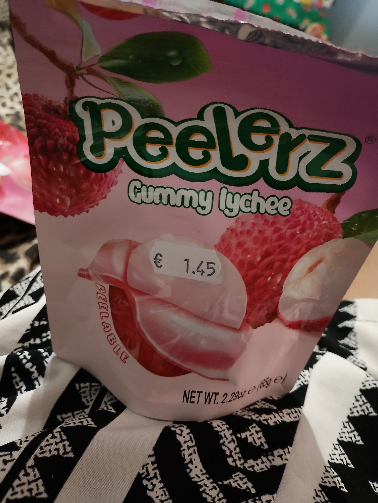 The image shows a package of "Peerlerz Gummy Lychee" candy. The packaging is predominantly pink with green and orange text. There are images of lychees on the front. A price tag with "€ 1.45" is attached to the packaging, and the words "PEELABLE" are visible vertically on the left side. The bottom of the package shows "NET WT. 2.29oz e (65g e)". The package is lying on a black and white striped fabric. There is no identifiable information about the location in Zimnitsa, Bulgaria within the image itself.
