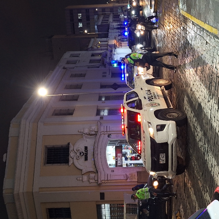 A white police truck with flashing red lights is parked on a cobblestone street. Police officers in neon green vests are seen nearby. The truck has "POLICIA COMISARIA MIRAFLORES" printed on it. A building with a sign "WALON" is in the background. Streetlights illuminate the scene at night. Another police motorcycle with blue lights is further down the street.