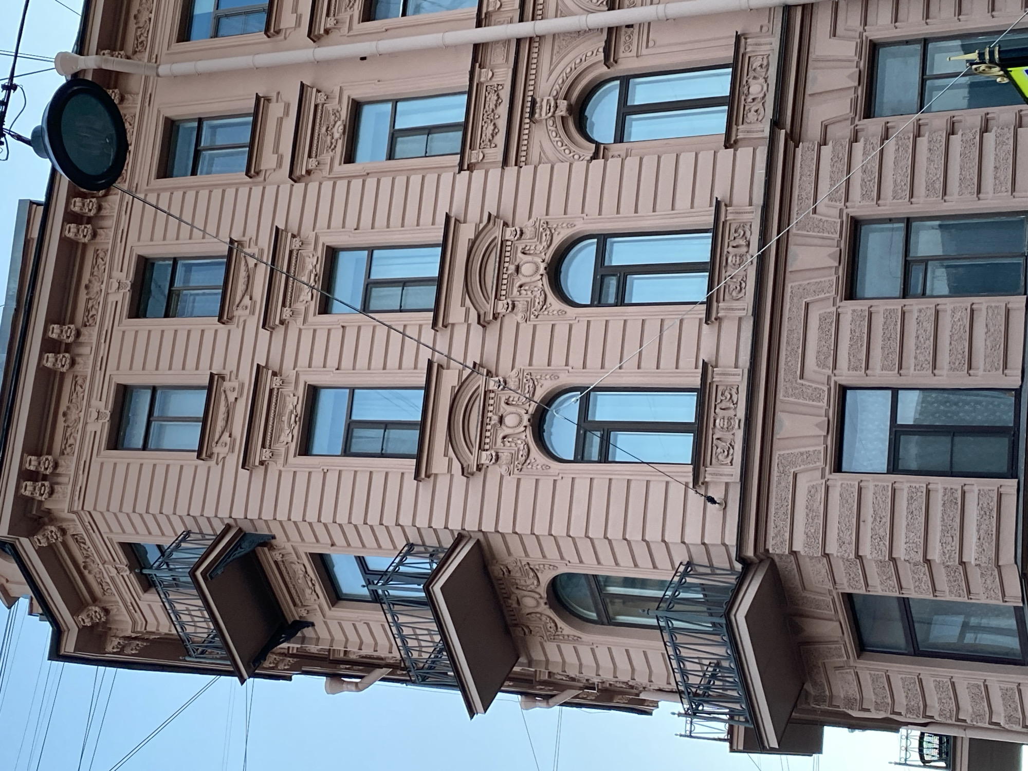 The image shows a light-colored building facade in Saint Petersburg, Russia. The building features ornate architectural details, including patterned columns, decorative window frames, and arched windows. Some windows have small balconies projecting outwards. There is a streetlight attached to the building. Electrical wires are visible against the pale blue sky.