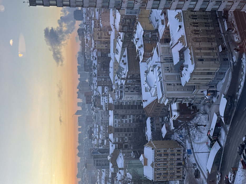 The image depicts a winter cityscape in Kyiv, Ukraine, observed from an elevated vantage point, likely during dawn or dusk, indicated by the orange-pink gradient at the horizon. Snow uniformly covers all rooftops, ground surfaces, and tree branches, with streets partially cleared for traffic. In the mid-ground, the prominent light-colored, ornate building with arched windows is identifiable as the Brodsky Synagogue (Central Synagogue), situated near Shota Rustaveli Street. To its left, a distinct multi-story yellow building with decorative facade elements stands, while various residential blocks, ranging from older ornate structures to more functional multi-story buildings, fill the scene. A main street occupies the lower left foreground, where several vehicles are parked or in transit, and a few pedestrians are visible on snow-ddusted sidewalks. Commercial kiosks or stalls line parts of the street. In the distant background, numerous high-rise buildings create a hazy skyline. A distinct plume of smoke or steam rises into the sky in the upper right background. Reflections on the upper portion of the image suggest it was taken through a window.