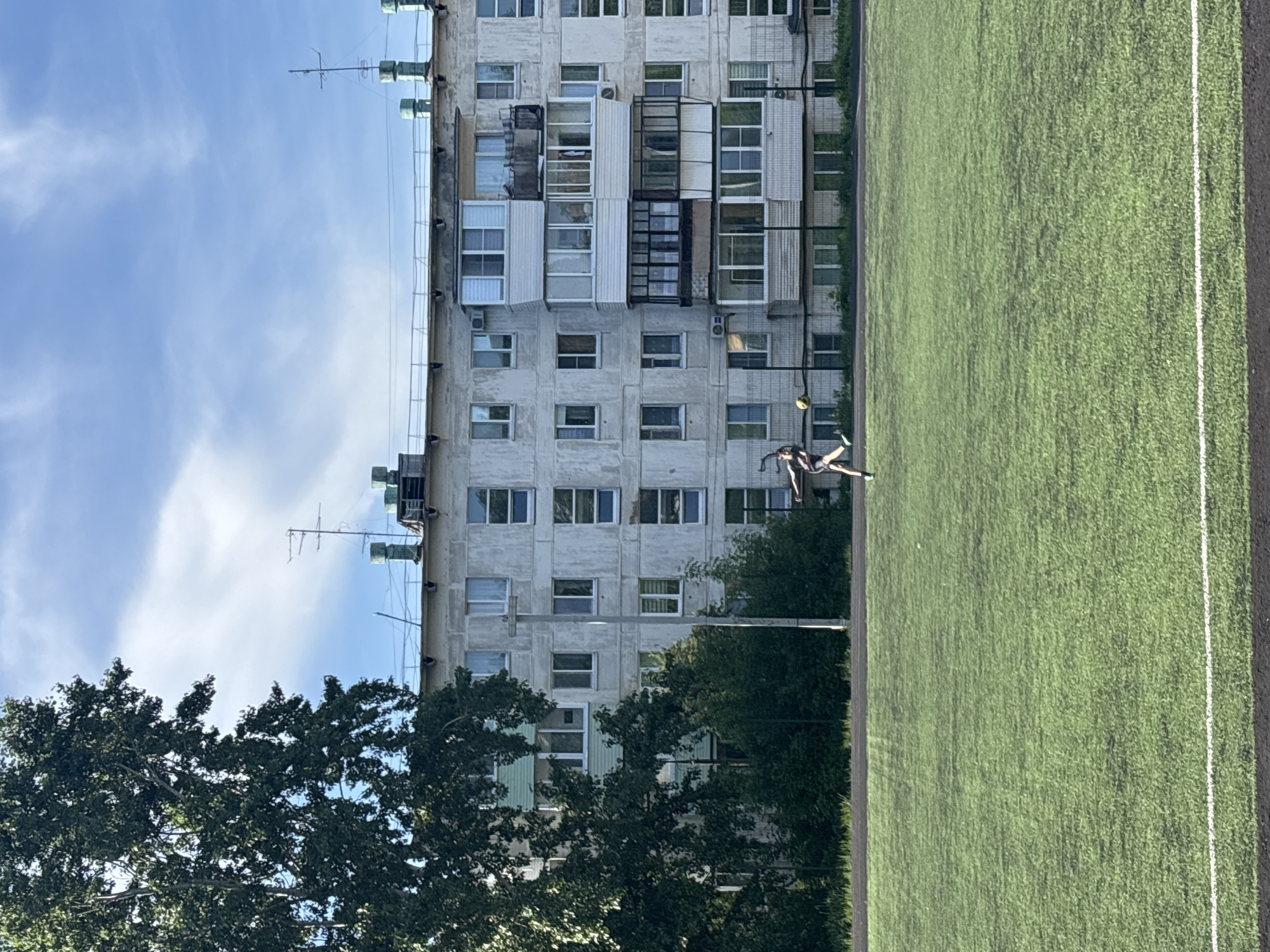 The image depicts a soccer field adjacent to a multi-story apartment building under a blue sky with scattered clouds. In the foreground is a section of green artificial turf soccer field with a white line marking a boundary. A person is kicking a soccer ball, mid-action, on the field. Trees obscure the base of the building. The apartment building is a gray, rectangular structure with numerous windows and enclosed balconies. Antennas are visible on the roof. The location is Chaikovsky, Russia.