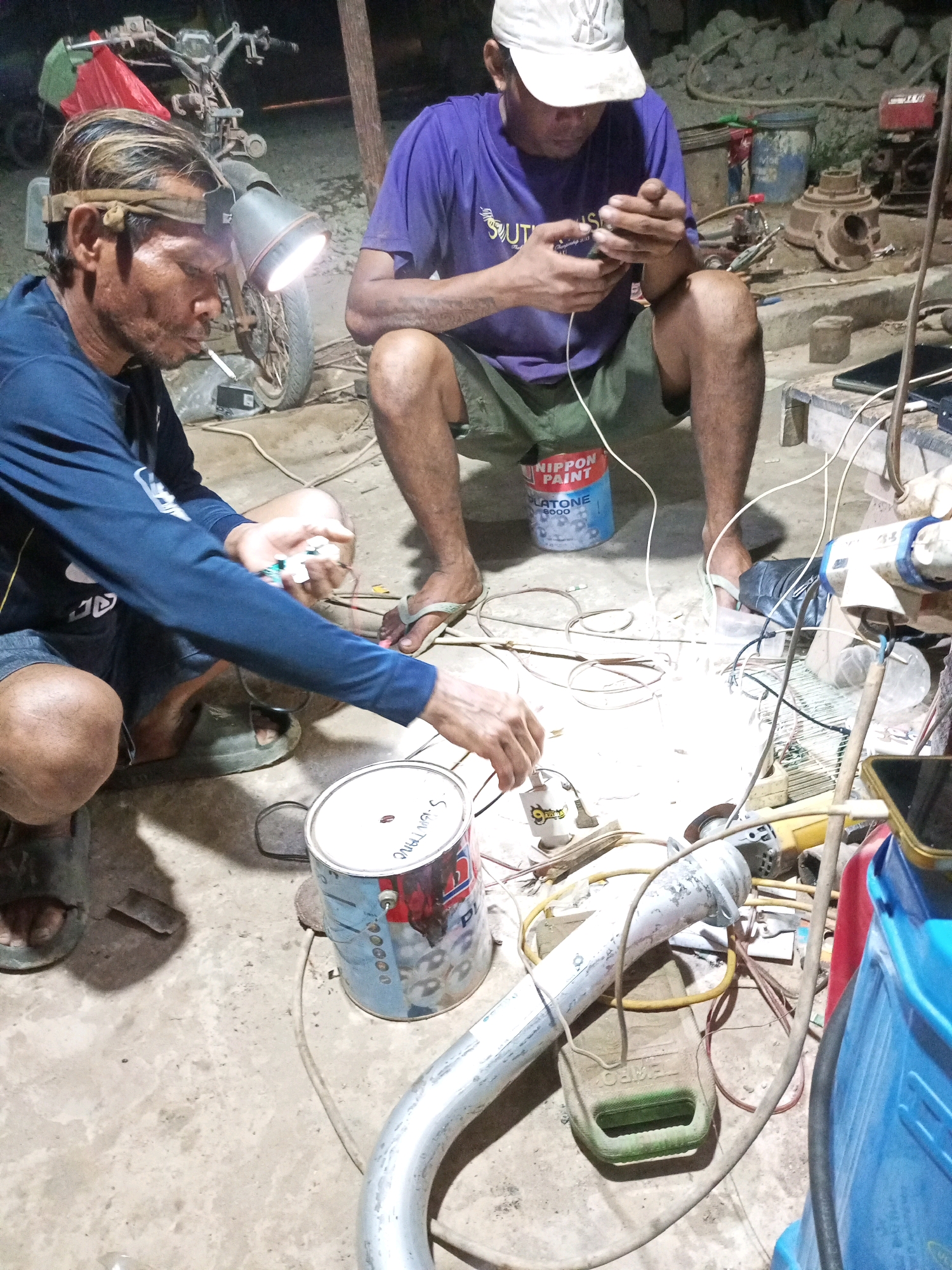 The image depicts an outdoor scene at night, likely in Keling Panggau, Indonesia, with two men engaged in electrical work amidst a cluttered environment of tools and discarded materials. The foreground shows a man on the left wearing a dark blue long-sleeved shirt and dark shorts, with a headlamp illuminating his face and work area. He is holding a cigarette and appears to be connecting wires. To his right, another man, wearing a purple t-shirt and green shorts, sits on a red and blue paint bucket and is focused on a small electronic device in his hands, possibly a mobile phone. His white baseball cap is visible. The ground is concrete and scattered with various items including wires, a metal pipe, a grinder, plastic containers, and what appears to be a motorcycle in the background. The lighting is artificial and stark, casting long shadows. The overall impression is one of a makeshift workspace or repair area.