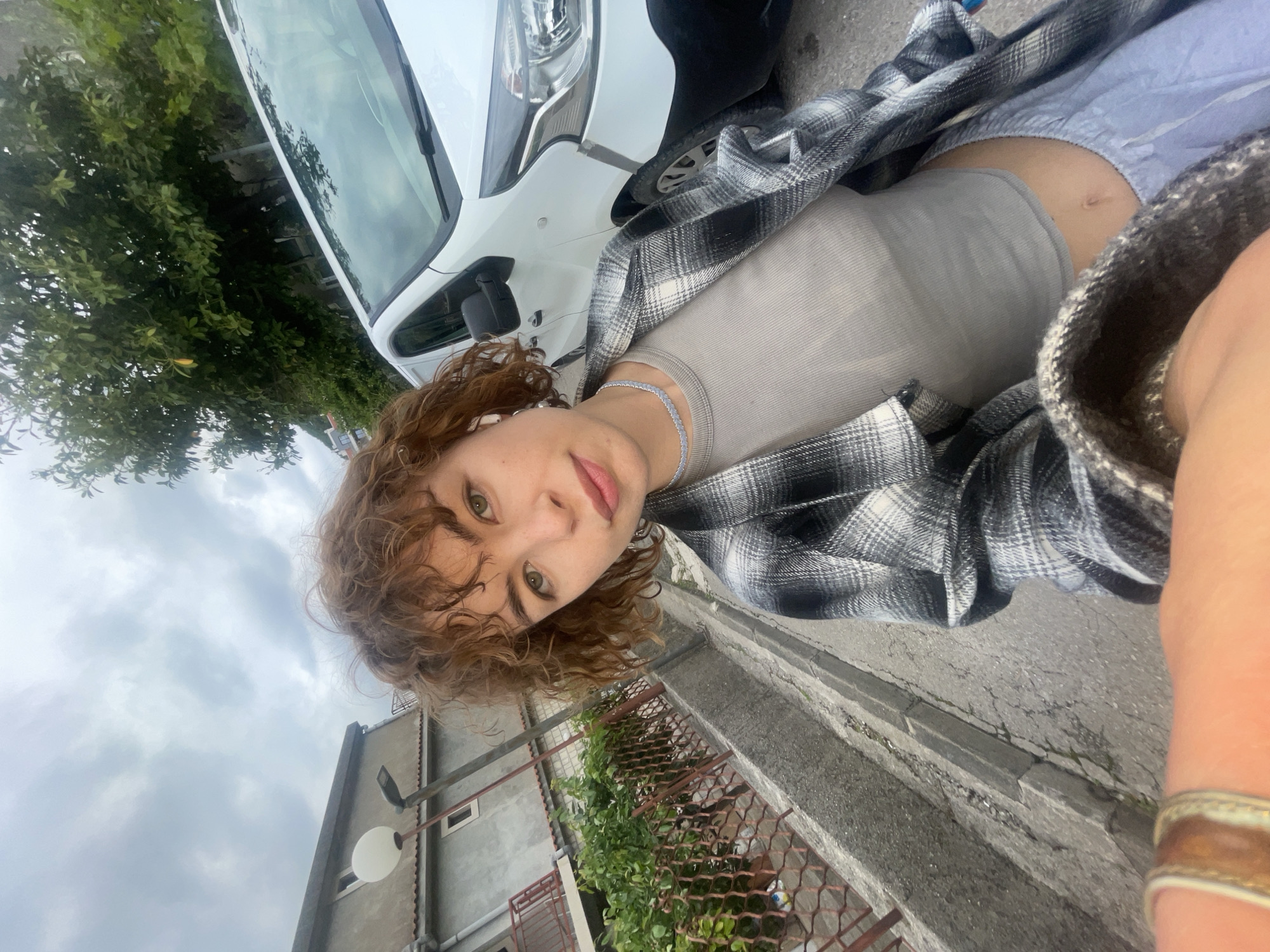 A young, fair-skinned person with curly reddish-brown hair is pictured in a selfie-style shot. They have green eyes and are wearing a light grey top, a silver choker necklace, and a black and white plaid flannel shirt. The background includes a white van, trees, a building with a red iron fence and greenery, and a paved street with cracked asphalt. The image may have been taken in Budva, Montenegro, although there are no immediate elements visible to confirm the exact location.