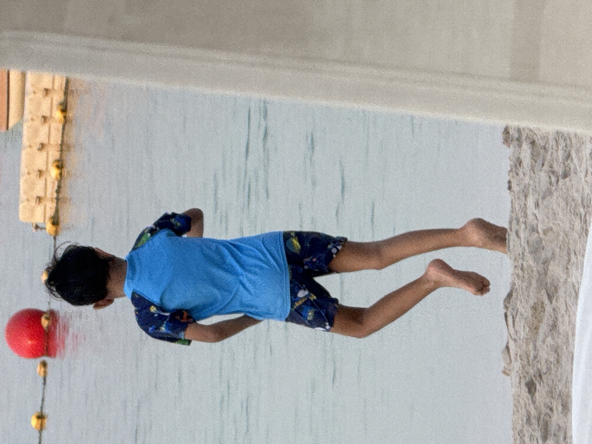 A young boy wearing a blue rash guard with a colorful pattern on the sleeves and dark swim trunks with a similar pattern is captured from behind, facing a body of water. He is standing on a sandy beach. In the background, a large red ball floats in the water, marked by buoys and a rope. Further in the distance, a structure resembling a dock or platform is visible with yellow buoys. The overall environment appears to be a beach area in the United Arab Emirates.