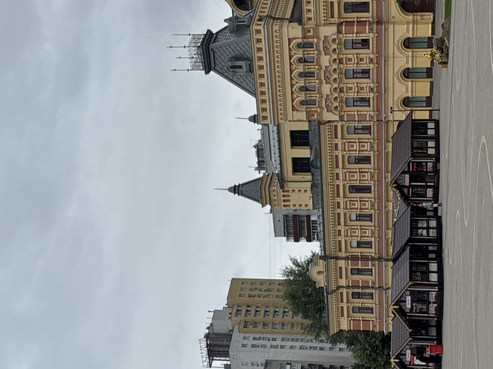 The image captures a view of the Nizhny Novgorod Kremlin, specifically the Merchant House (Gostiny Dvor), under an overcast sky. The foreground features a paved square with several small, dark-roofed market stalls lined up. A few individuals are visible; one person stands by a red bin near the left stall, and another person in a checkered shirt walks near the right side of the stall arrangement. A decorative display of sunflowers and rocks sits in the middle ground to the right of the stalls. The main subject is the ornate yellow and red building of the Merchant House, characterized by its detailed facade, numerous arched windows, and a prominent dark roof with decorative spires. To the left of the Merchant House, a tall, modern apartment building with a light-colored facade and numerous windows stands out against the historical architecture. The environment appears to be an urban setting, likely a public space within the city of Nizhny Novgorod, Russia.