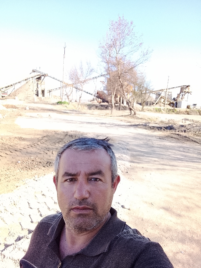 An adult male is prominently featured in the foreground of a daylight selfie, positioned in Yangi Chinoz, Uzbekistan. He has graying dark hair, a short beard, and a neutral expression, looking directly into the camera. He is wearing a dark brown collared shirt with horizontal stripe patterns on the shoulders. The background reveals an industrial or construction environment characterized by light brown, uneven ground with visible earth disturbances. Multiple industrial structures dominate the mid-ground, including several interconnected conveyor belt systems supported by metal frameworks, leading to and from large aggregate piles and processing machinery. A tall, slender pole stands vertically near the center of the background. Scattered leafless deciduous trees are present throughout the scene. The sky is clear and light blue, indicating fair weather conditions.