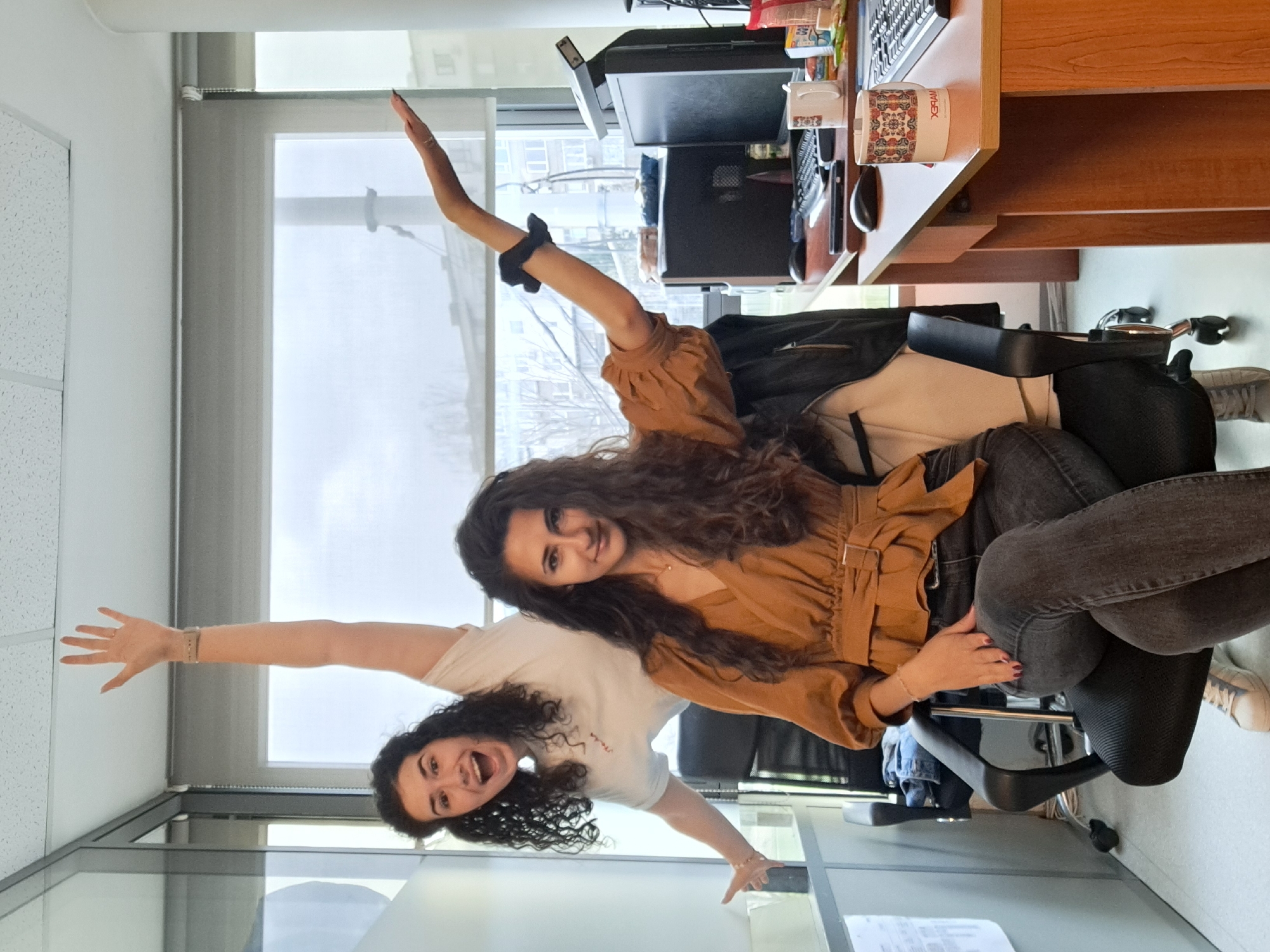 Two women are posing in an office. One woman with curly brown hair sits in an office chair, wearing a brown, button-down shirt and dark jeans. She raises one arm. Behind her stands another woman wearing a white t-shirt with curly dark hair. She has both arms outstretched. The office has a window with a view of a city, a desk with a computer and other office supplies, and a glass partition. Based on the architecture visible through the window and interior of the office, this image likely captured in Sofia, Bulgaria.