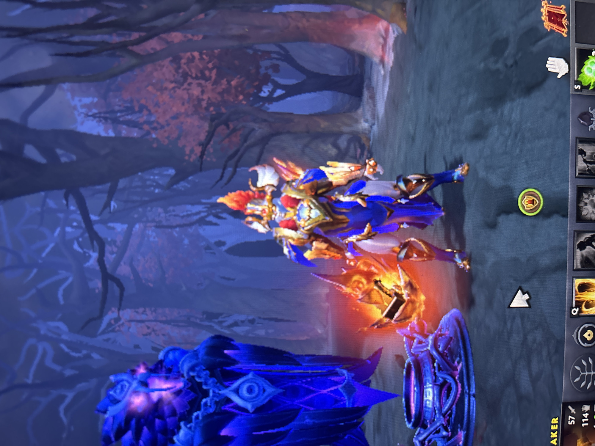 A digital screenshot from a video game depicts a fantastical scene, rotated 90 degrees clockwise. In the center, a character, possibly a hero or knight, stands in ornate blue and gold armor with prominent orange and red accents. The character emits a bright orange glow, suggesting magical energy or fire. To the left of the character, a large, dark blue, organic-looking structure with glowing purple elements and an eye symbol is visible, possibly a defensive tower or siege unit. Part of another similar dark blue/purple structure is to the right of this main structure.
The background features a forest with dark, gnarled tree branches and sparse reddish-orange foliage, casting a mystical atmosphere. The lighting suggests either dusk or night, with deep blue and purple tones dominating the environment.
On the right side of the screen, a user interface (UI) is visible. It includes various icons arranged vertically: a red framed picture icon at the top, followed by a white hand icon, and a bright green, spiky, glowing icon with the letter "S". Below these are several other unlit or greyed-out ability/item icons, including a circular gold icon with a shield emblem. A white arrow-shaped cursor points towards the lower-middle right. At the very bottom right, text "AKER" (likely part of a character or item name) is partially visible, alongside numbers "57" and "114" with small associated icons, possibly indicating in-game resources or stats. The scene captures an in-game moment, likely during gameplay or a strategic stand-off.