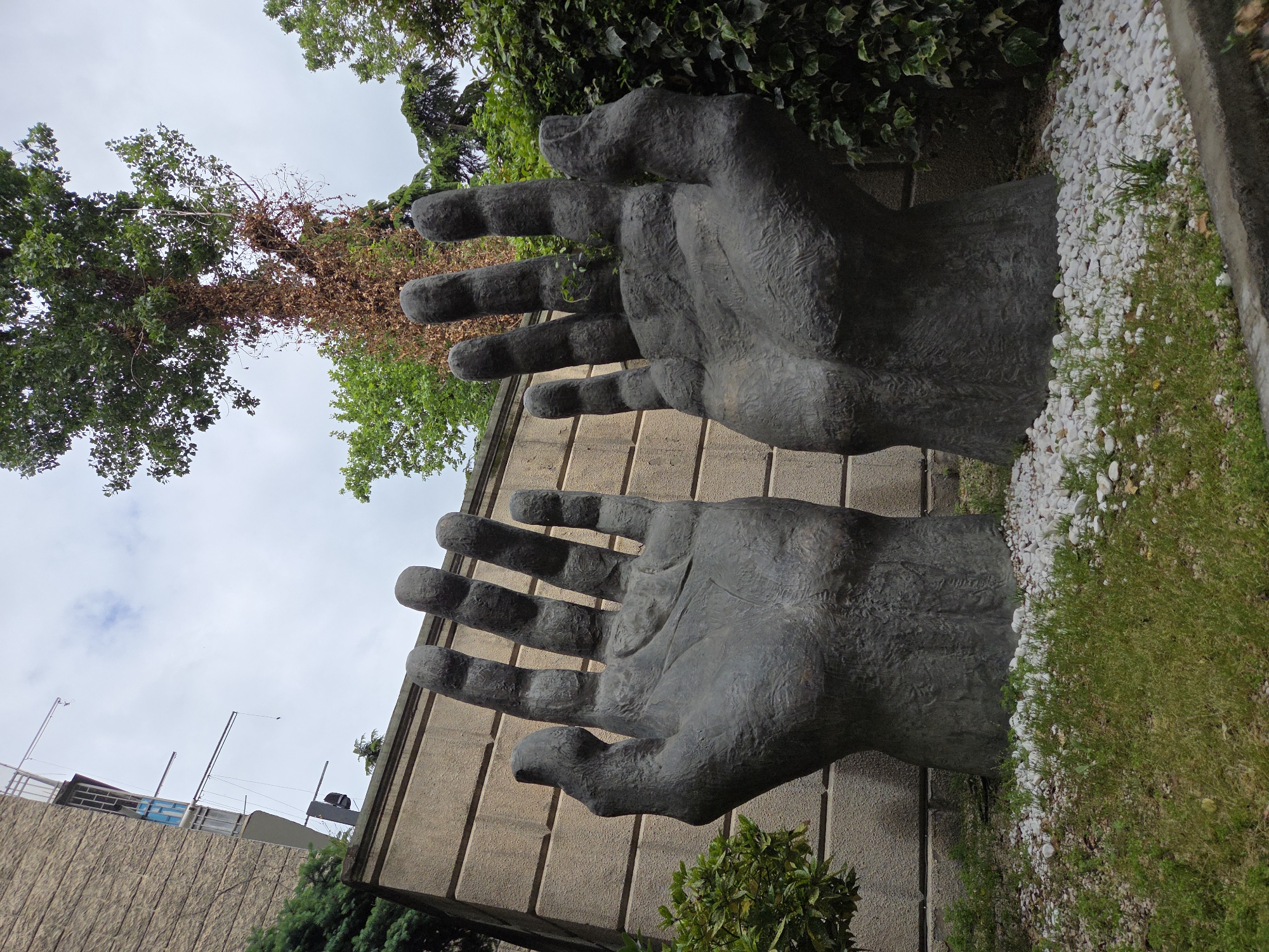 The image features a pair of large, dark-colored, sculpted hands seemingly emerging from the side of a building. The hands are open, with fingers extended, and have a textured, slightly weathered appearance. The building has a façade of vertical panels. Adjacent to the base of the hands is a strip of grass and small white rocks or pebbles. The sky is overcast, visible between the hands and around the surrounding trees and foliage. A modern building structure can be seen in the background, alongside some power lines. The location is the Hands of Peace in Unknown, Unknown.