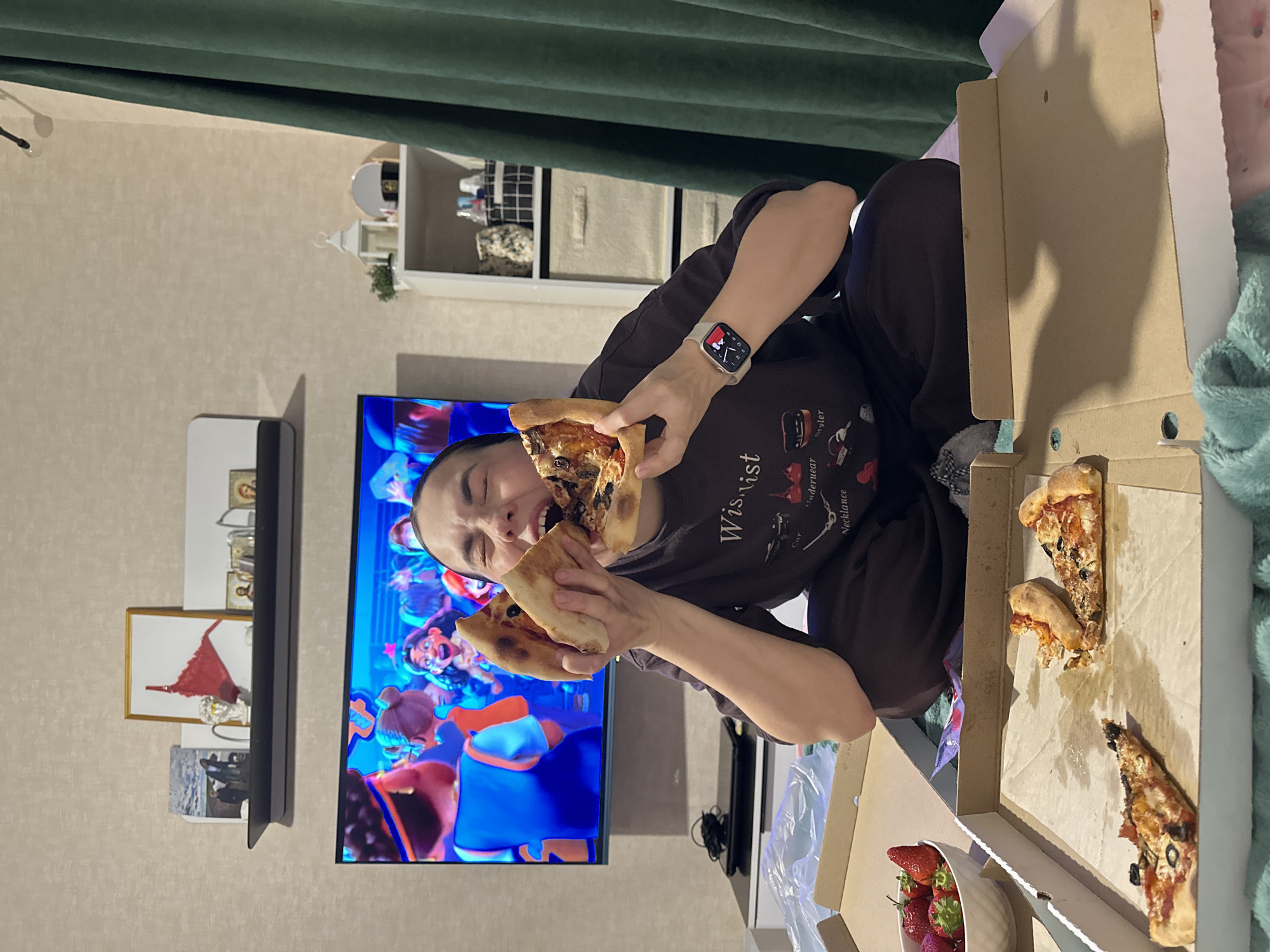 A person is eating two slices of pizza in front of a television, possibly in a home in Perm, Russia. The person is wearing a dark-colored sweatshirt with writing on it and a smartwatch. They have a bowl of strawberries nearby, and the pizza is in an open cardboard box. The TV screen displays an animated image. A shelf with decorative items is visible on the wall. A green curtain is in the background.