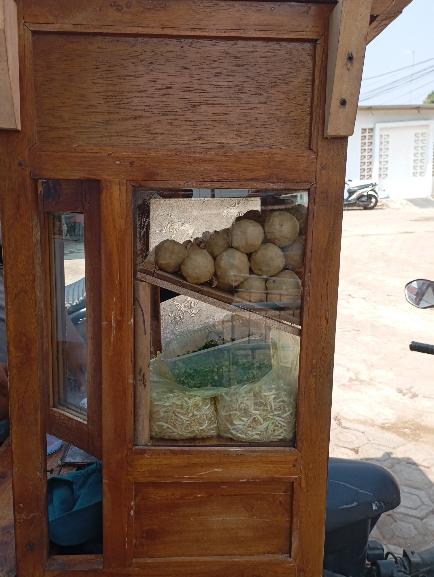 An outdoor, eye-level, medium shot captures a wooden food cart with glass panels displaying food items. The cart's wood is a medium brown, showing a visible grain and some wear. Inside the cart, stacked on a wooden shelf, are approximately ten round, light brown food items, likely meatballs, some of which are partially obscured by a decorative metallic backing. Below the meatballs, two clear plastic bags contain ingredients. The bag on the left holds finely chopped green vegetables, possibly scallions or herbs. The bag on the right is filled with light-colored bean sprouts. The cart is positioned on a paved surface, possibly a sidewalk or road, with the background showing a white building with a repeating pattern of small square openings and a white fence. A motorcycle is parked to the right of the cart, partially visible. The sky is clear and bright. The scene appears to be in an urban or semi-urban environment.