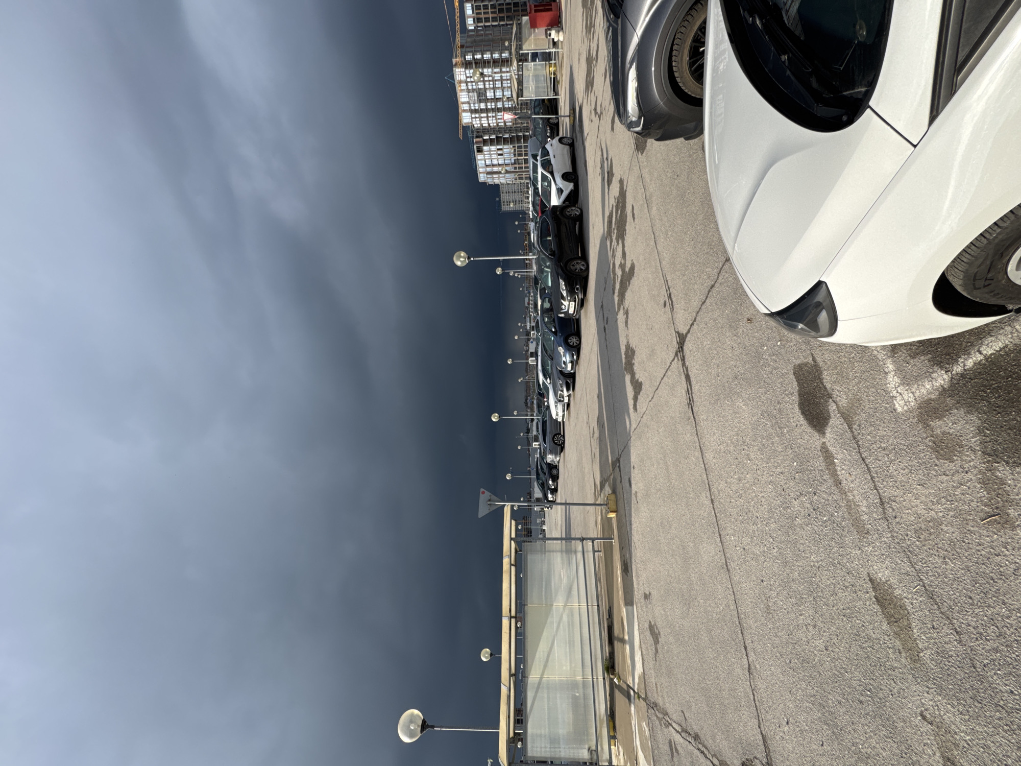 The image depicts a parking lot in Burgas, Bulgaria, filled with cars. A row of streetlights extends along the parking area. In the background, a building under construction is visible. The sky is dark and overcast, suggesting an approaching storm. The foreground features a white car and parts of other vehicles. There is a bus stop enclosure along the parking lot, with a sign visible next to it. The pavement shows signs of wear and water stains.