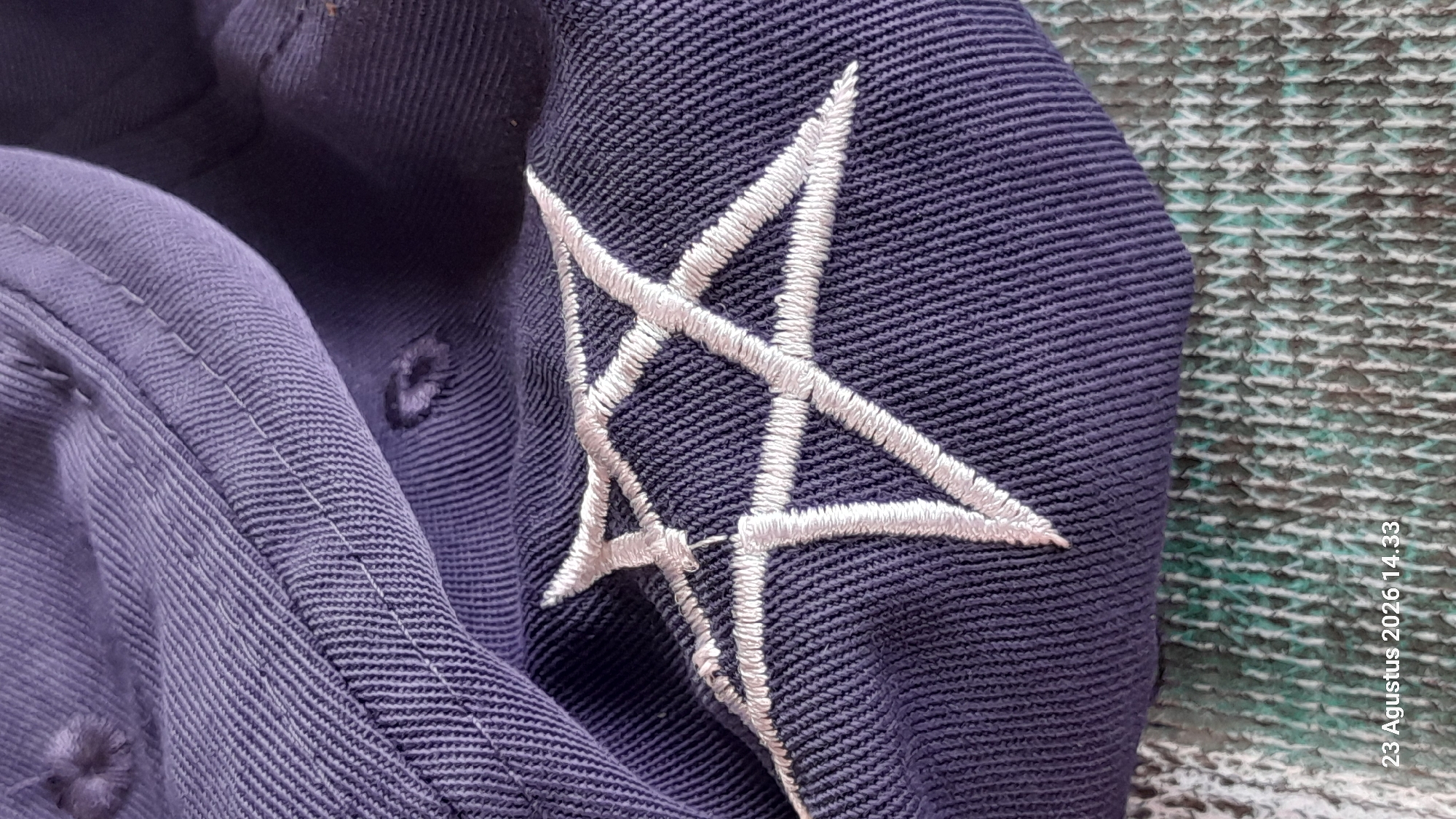 This is a close-up, flat lay image of a dark purple baseball cap. The cap has white embroidery in the shape of a star, formed by five connected lines. The fabric of the cap has a visible ribbed texture, and there are some small holes or frayed areas in the fabric. The background is a textured surface with a greenish-grey and white pattern. A timestamp "23 Agustus 2026 14.33" is visible in the bottom left corner of the image. The image does not depict any specific location within the city of Gemaharjo, Indonesia, as it focuses solely on the object itself.