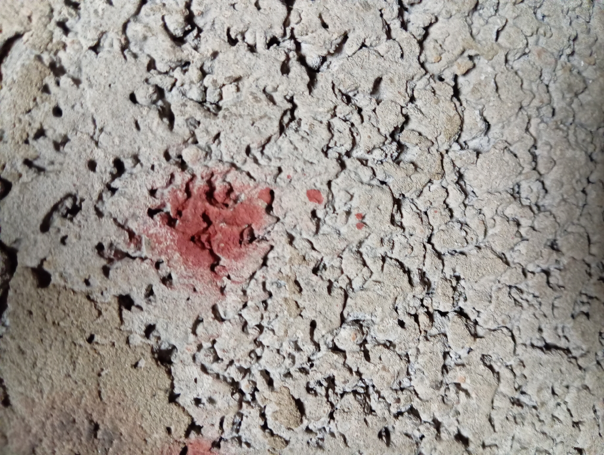The image shows a close-up view of a rough, porous concrete surface with red spray paint marks. The concrete has an uneven texture with many small holes and crevices. There are two visible red paint marks. One is a larger, more amorphous splotch located slightly above the center of the image. Below and to the left of this larger mark, there are smaller, scattered red paint dots. The lighting suggests the surface is outdoors, possibly a wall or a construction material. The location in Hlynytsi, Ukraine, is noted, but no specific environmental or urban context beyond the concrete surface itself is discernible from the image.