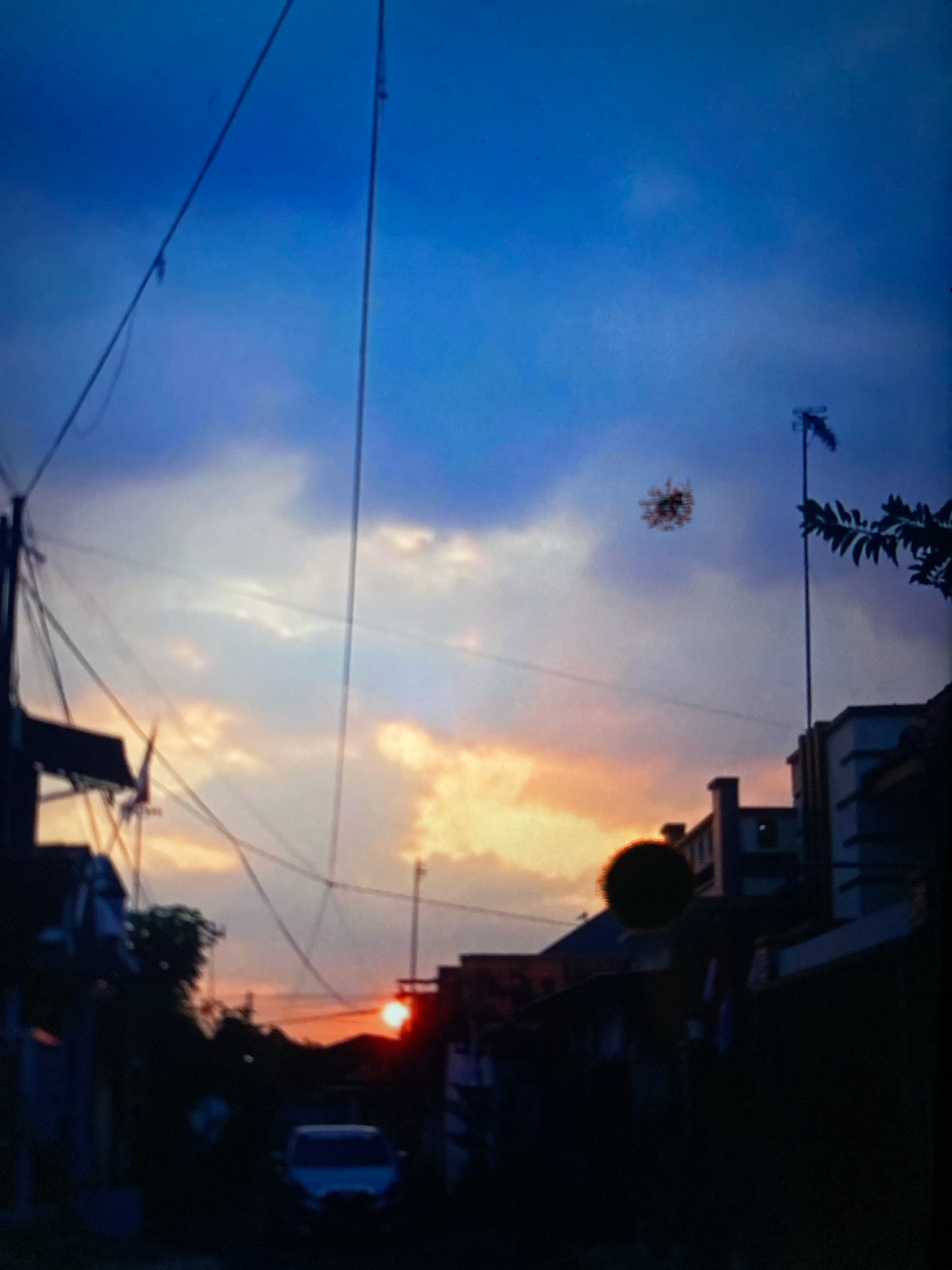 The image depicts a sunset over a city street in Probolinggo, Indonesia. The sky is a gradient of deep blue, purple, and orange, with scattered clouds illuminated by the setting sun. The sun itself is a bright orange orb visible near the horizon, partially obscured by buildings. Silhouetted against the sky are power lines, telephone poles, and the rooftops of buildings. In the foreground, a street is visible, with a white van parked on the right side. The overall impression is one of a calm, urban evening.