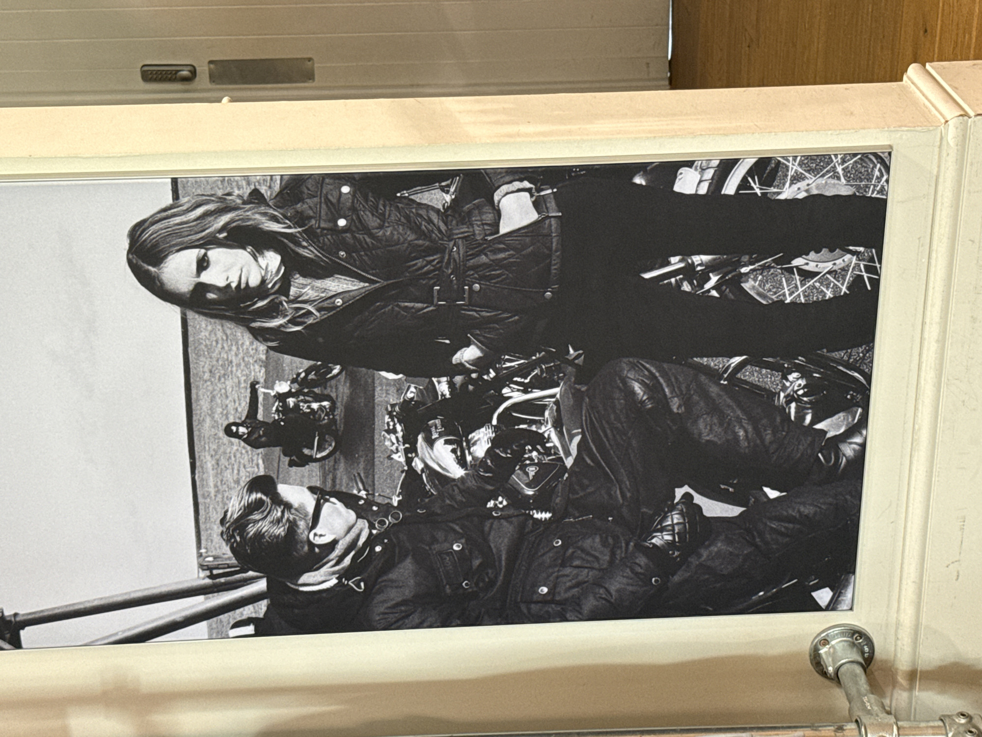 This is a black and white photograph of a fashion advertisement featuring two individuals posing with motorcycles. The woman on the right is looking directly at the camera, wearing a quilted jacket and dark pants. The man on the left is seated on a motorcycle, looking to the side, also dressed in a jacket and pants. In the background, another person is riding a motorcycle. The photograph is framed by a cream-colored border. The photograph appears to be displayed in a store or public space, indicated by the visible door handle and lock mechanism on the right side of the image and a metal pipe fixture on the left. The setting appears to be outdoors near water. The image is likely a poster or a print displayed for commercial purposes.