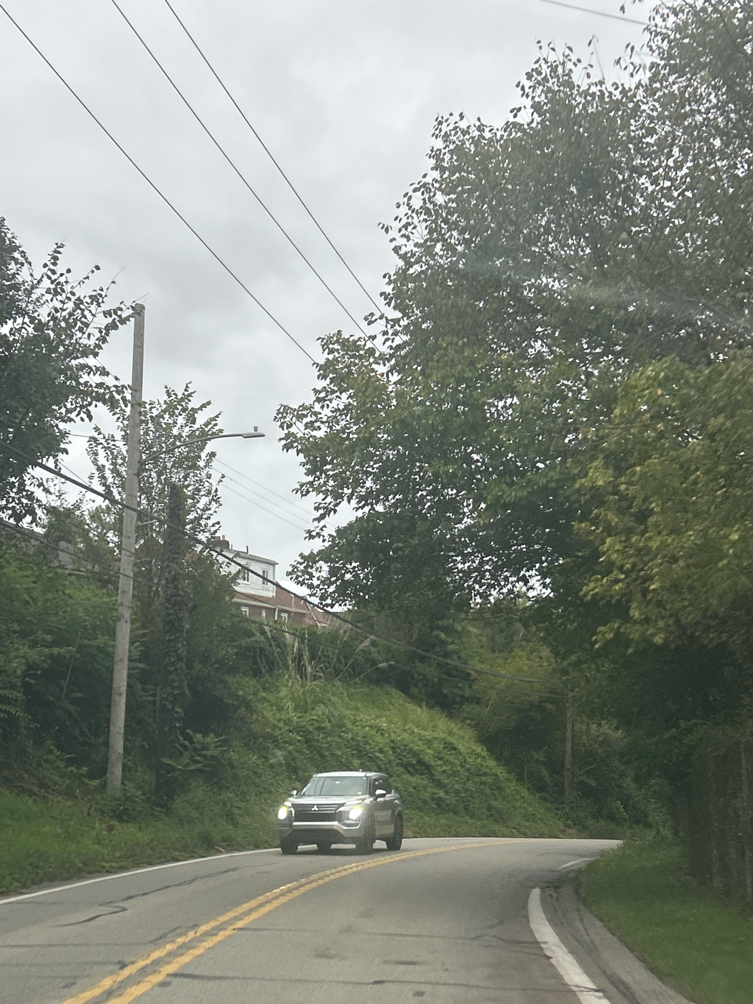 The image captures a frontal view of a silver SUV driving on a two-lane asphalt road. The road has a yellow double line in the center and a white line on the right edge. The SUV is heading towards the viewer, slightly to the right of the road's center. The surrounding environment is characterized by lush green vegetation, including trees and bushes, flanking the road. A utility pole with wires is visible on the left side of the road, partially obscured by trees. In the background, a large white building with a red-tiled roof is situated on a grassy slope. The sky is overcast, indicating a cloudy day. The scene appears to be a residential area with natural elements, possibly located in Carnegie, United States of America, given the context.