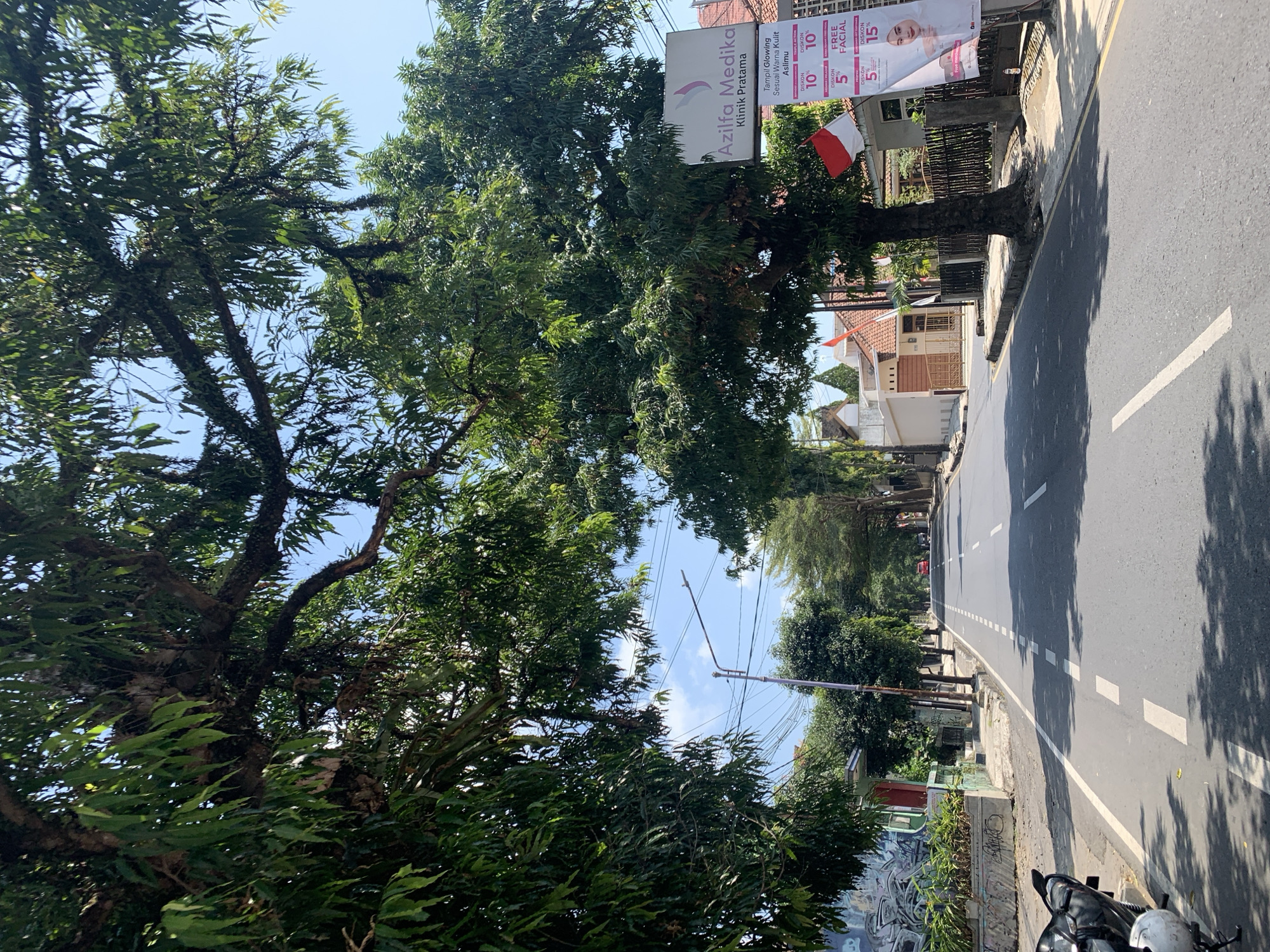 The image captures a street scene in Ciamis, Indonesia, under a clear blue sky. Large, leafy trees arch over the road, casting dappled shadows on the asphalt. The road itself is paved and marked with white dashed lines. On the left side of the road, there's a building with graffiti art and a red door. Further down, a street lamp and power lines are visible. On the right side, a sign for "Azilfa Medika Klinik Pratama" is displayed, featuring promotional offers like "DISKON 10%" and "FREE FACIAL." A small Indonesian flag is mounted near the sign. Houses with brown fences line the street, and several trees are planted along the sidewalk. In the foreground on the left, a black motorcycle is parked. A red car is seen in the distance, further down the road. The overall impression is of a quiet, sun-drenched street in a residential or commercial area.