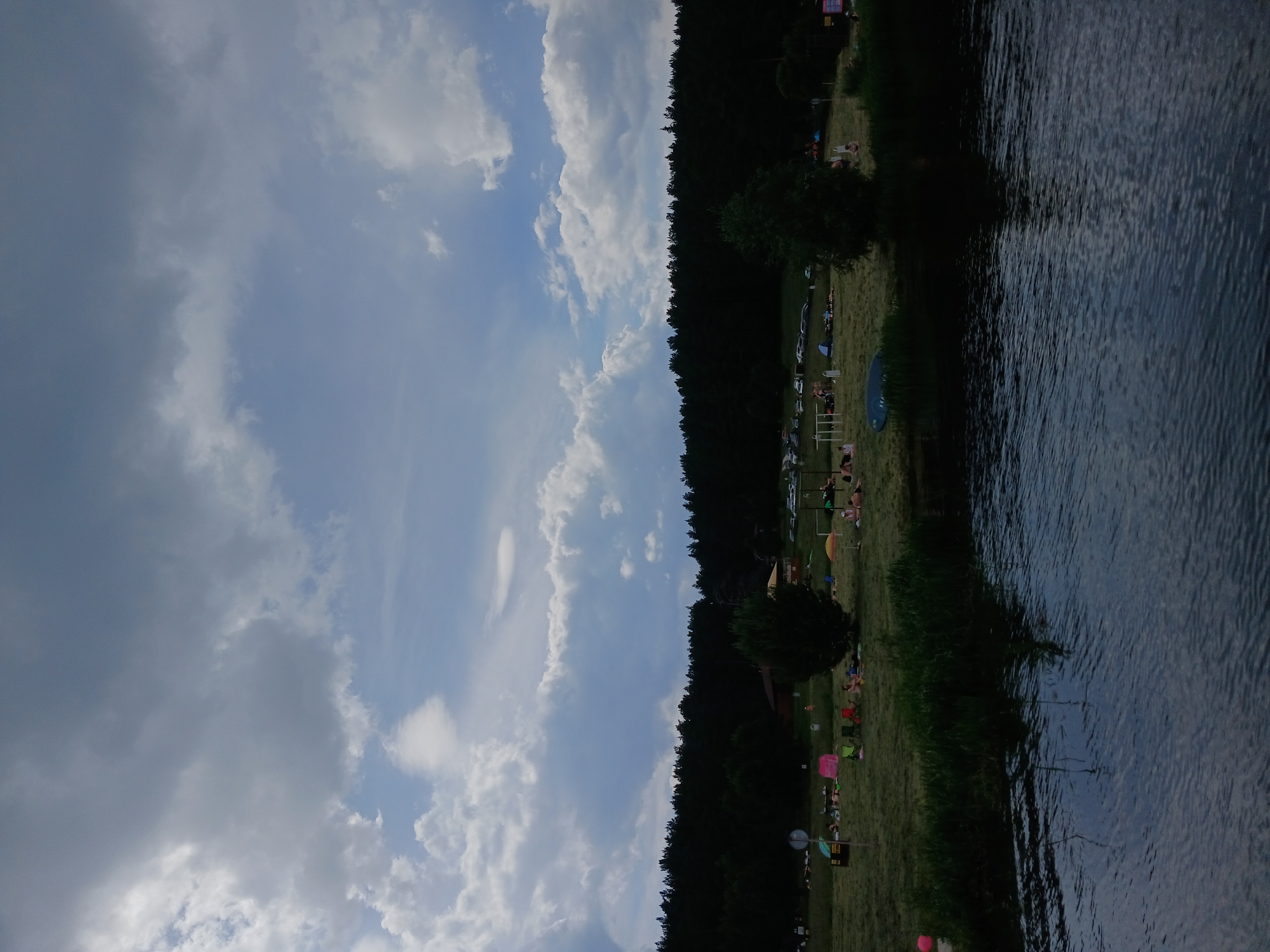 The image captures a daytime scene at a lakeside recreational area, identified as being in the city of Revda, Russia. The foreground features a body of water with ripples on its surface. The middle ground shows a grassy bank dotted with people sunbathing, sitting on chairs, and relaxing under umbrellas and tents. Various objects like beach balls, bags, and a blue kayak are visible on the grass. Several cars are parked in the background, beyond the grassy area, adjacent to a dense line of dark green trees that forms the backdrop. The sky is partially cloudy, with a mix of dark and light gray clouds against a pale blue sky. A few buildings, possibly small structures or shelters, are partially obscured by the trees.