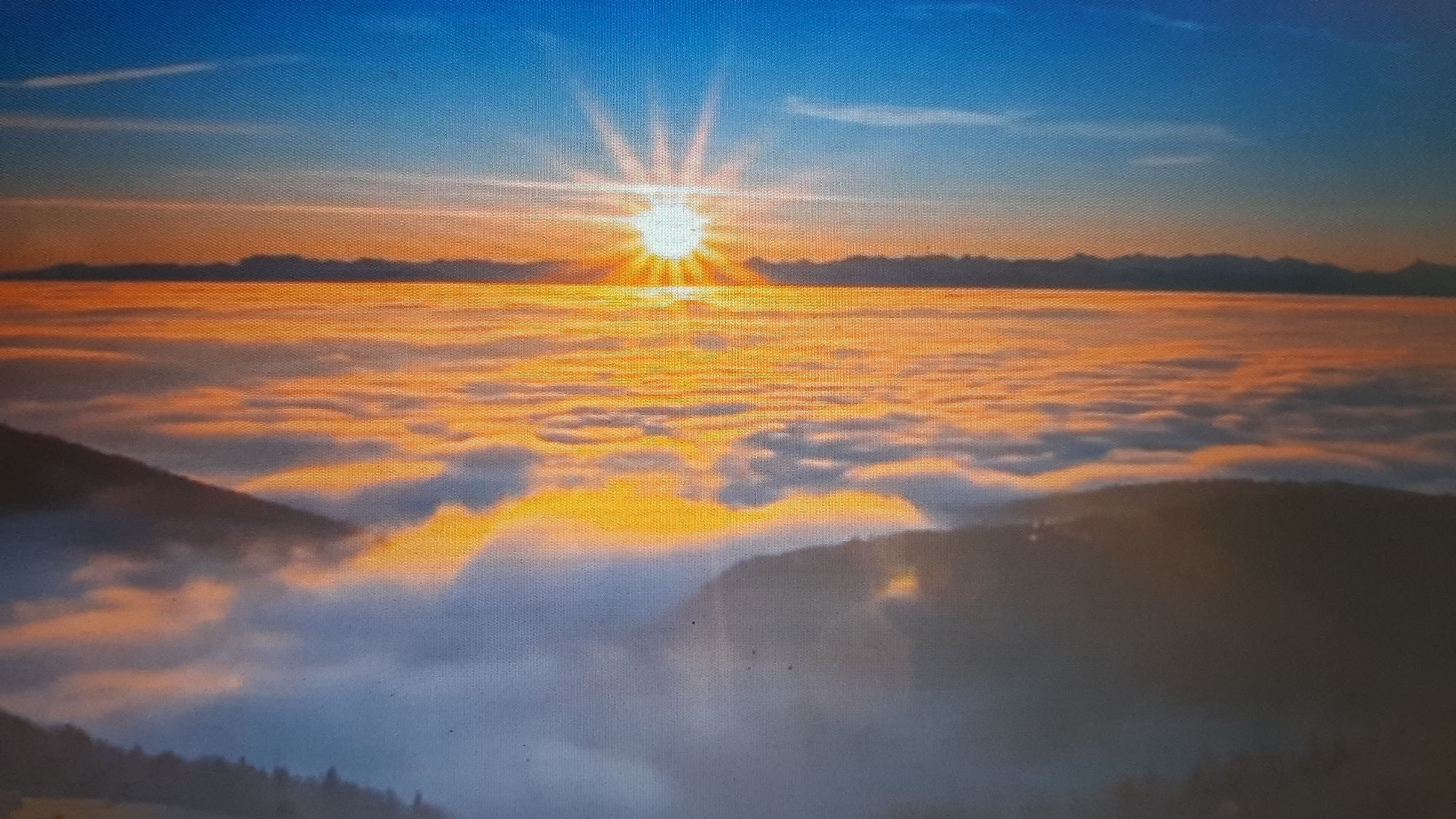 The image depicts a panoramic view of a sunrise over a sea of clouds, with the sun's rays radiating outwards. Mountains are visible in the distance, silhouetted against the bright sky. The foreground shows hills emerging from the clouds, covered in sparse trees. The scene is bathed in warm, golden light, suggesting early morning. The texture of the image appears to be a canvas print.