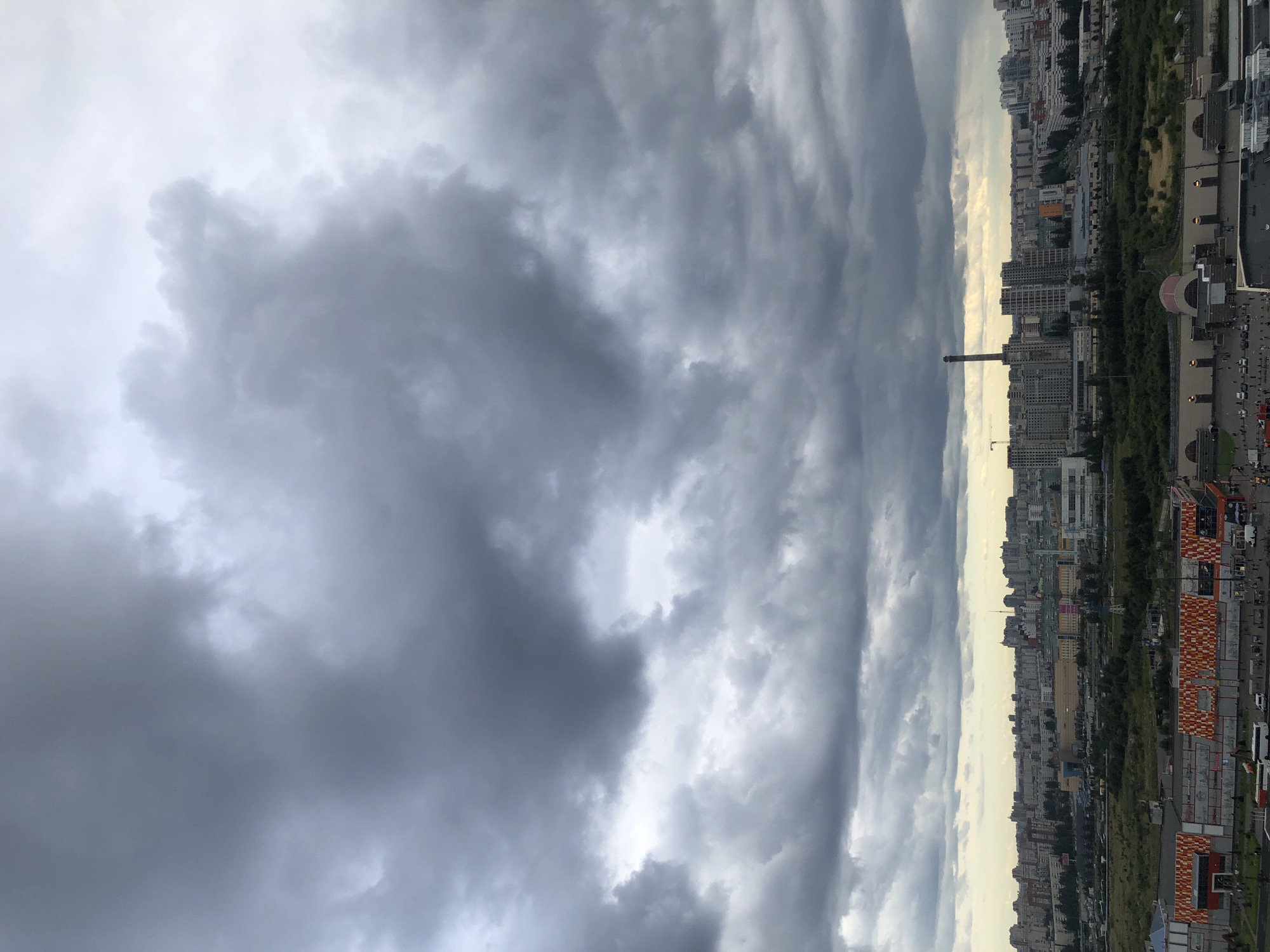 An eye-level, wide-angle shot captures a sprawling cityscape under a heavily overcast sky. The dominant feature is the dramatic cloud cover, a dense mass of dark, swirling grey formations that fill the upper two-thirds of the frame, obscuring any hint of blue. A faint, diffused light filters through the clouds, casting a muted glow over the urban landscape below.
The city itself appears to be densely populated, with a horizon dominated by a mix of residential and commercial buildings. The architecture suggests a modern urban environment, characterized by mid-rise to high-rise structures, predominantly in shades of grey, white, and beige. Some buildings feature visible windows, hinting at occupied spaces. In the middle ground, a tall, industrial chimney stands out, a solitary, dark vertical element against the urban backdrop.
In the foreground, the scene transitions to a commercial or industrial area. A large building with a distinctive facade, featuring a repeating pattern of orange and white rectangular elements, is prominent. Adjacent to this, a large parking area is visible, with several vehicles including buses and cars. Pedestrians are sparsely distributed, engaged in typical city activities. A band of green space, likely parkland or undeveloped land, separates the foreground structures from the denser city in the distance. The overall impression is one of a vast, modern metropolis under a looming, possibly stormy, sky.