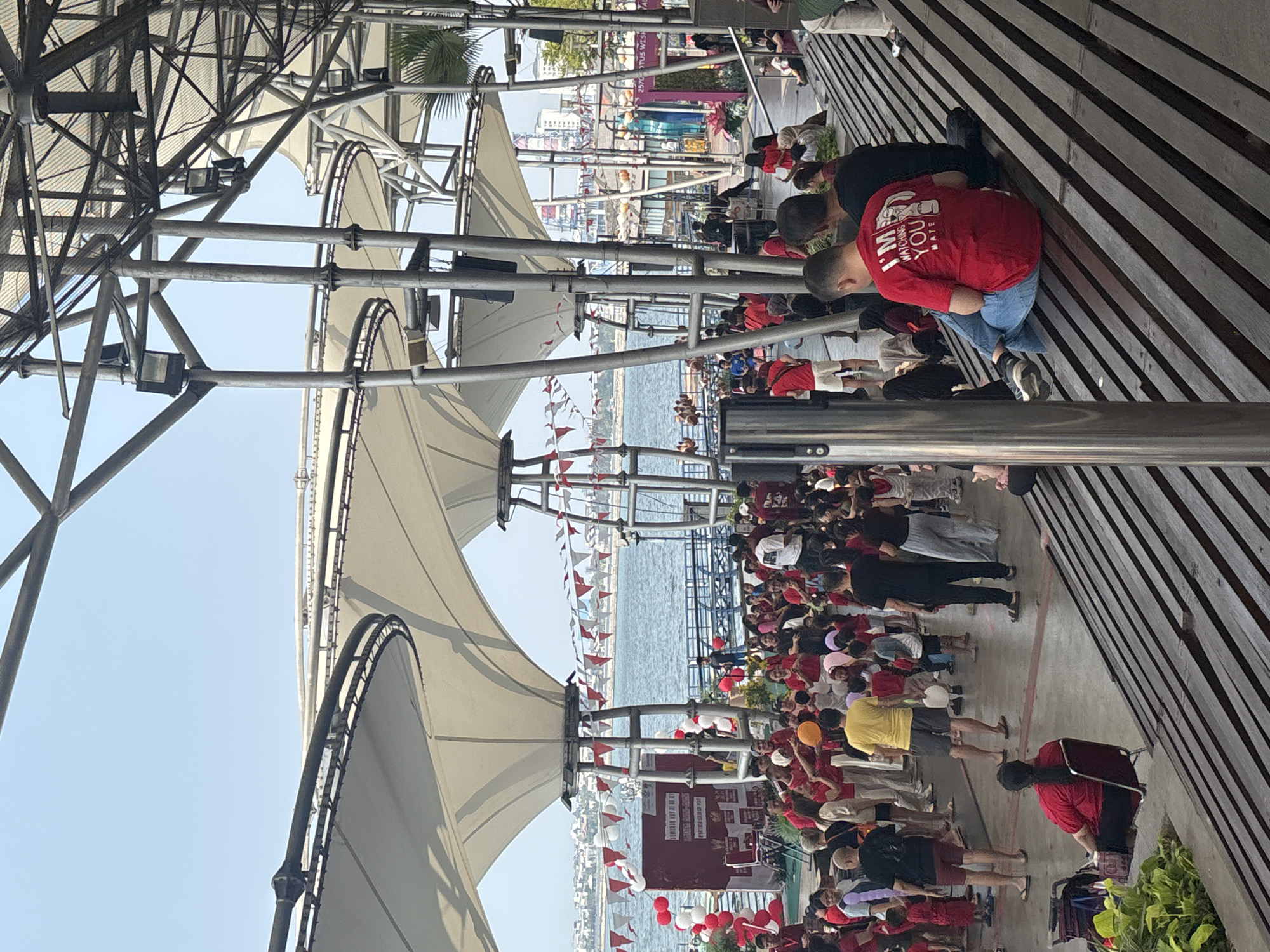The image depicts a public gathering outdoors in North Jakarta, Indonesia, likely at a waterfront location such as Ancol. A large crowd of people, including many children, are present under a modern, white-canopied structure supported by metal poles. Many individuals are wearing red attire, and red and white triangular flags are strung across the area, suggesting a festive or nationalistic event.
In the foreground, people are seated on wooden bleachers. One man in a red t-shirt with text that reads "I'M WATCHING YOU MATE" is prominently featured. Behind him, another man in a black t-shirt sits with his back to the camera. Other people are scattered throughout the scene, some standing and observing an activity or performance, while others are interacting with each other.
The background reveals a body of water, likely the Java Sea, with buildings and a cityscape visible on the horizon. The overall atmosphere appears to be lively and communal, with elements of a community event or celebration.