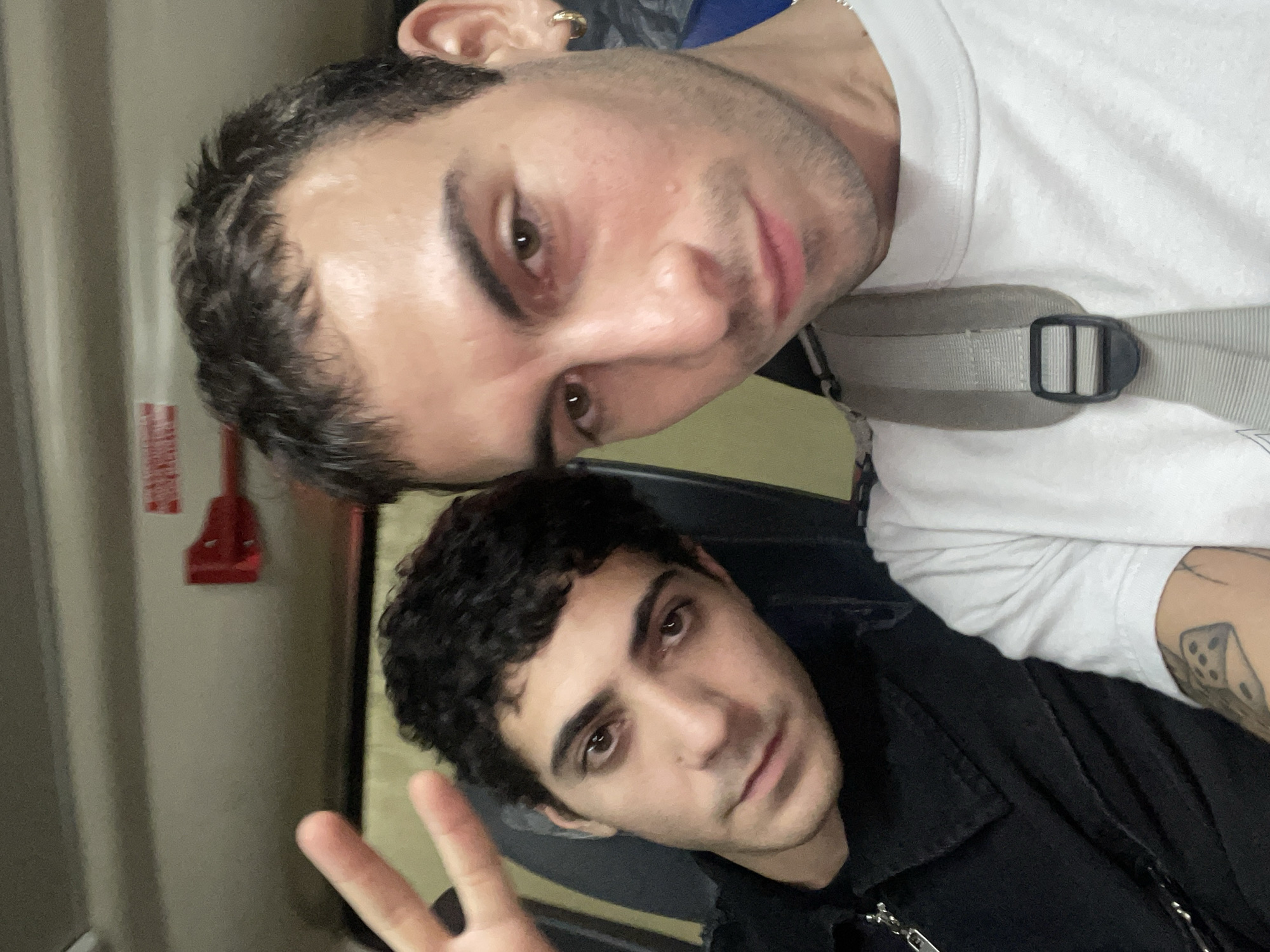 Two young men are featured in a selfie, likely taken indoors in Buenos Aires, Argentina. The man closer to the camera, positioned at the top, has dark hair, light stubble, and dark eyes. He is wearing a white t-shirt with a grey strap, possibly from a backpack or bag, draped across his chest. A gold-toned hoop earring is visible in his left ear. He looks directly at the camera with a relaxed expression.
The second man is positioned below and slightly behind the first. He has dark, curly hair and dark eyes, and is wearing a black jacket or shirt. He is making a peace sign with his right hand. A tattoo, possibly depicting dice or similar geometric shapes, is visible on his left forearm. He also looks towards the camera.
The background is softly out of focus, suggesting an indoor setting with neutral-colored walls, possibly grey or beige. A red object, which could be an emergency pull station or fire alarm, is mounted on the wall to the left of the second man. Faint, indecipherable white text is visible on this red object. The overall mood of the image is casual and friendly.