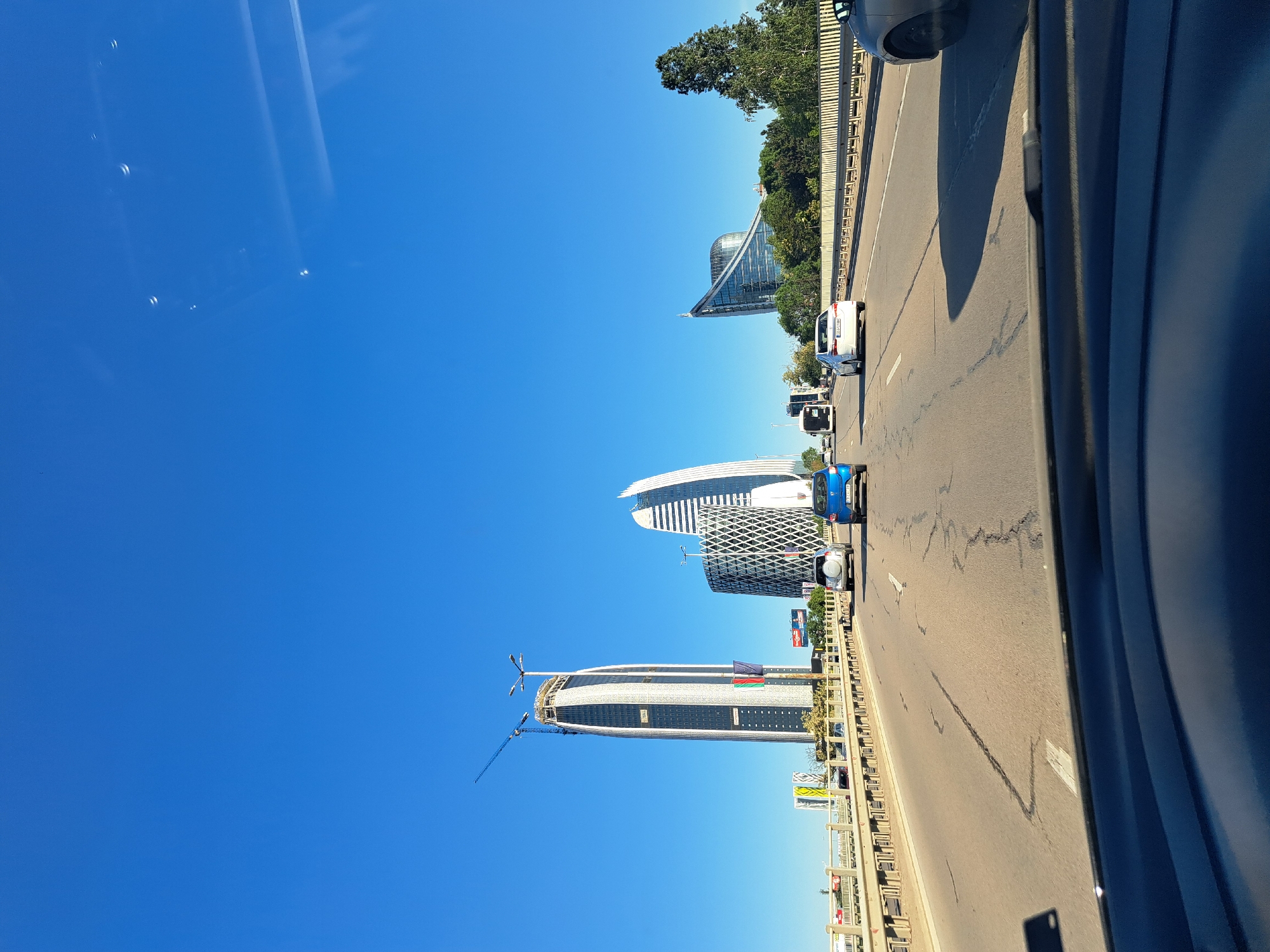 The image shows a street in the city of Unknown, Unknown. The street is lined with buildings, including a tall skyscraper under construction with a crane. There are cars driving on the street and a pedestrian bridge. The sky is blue.