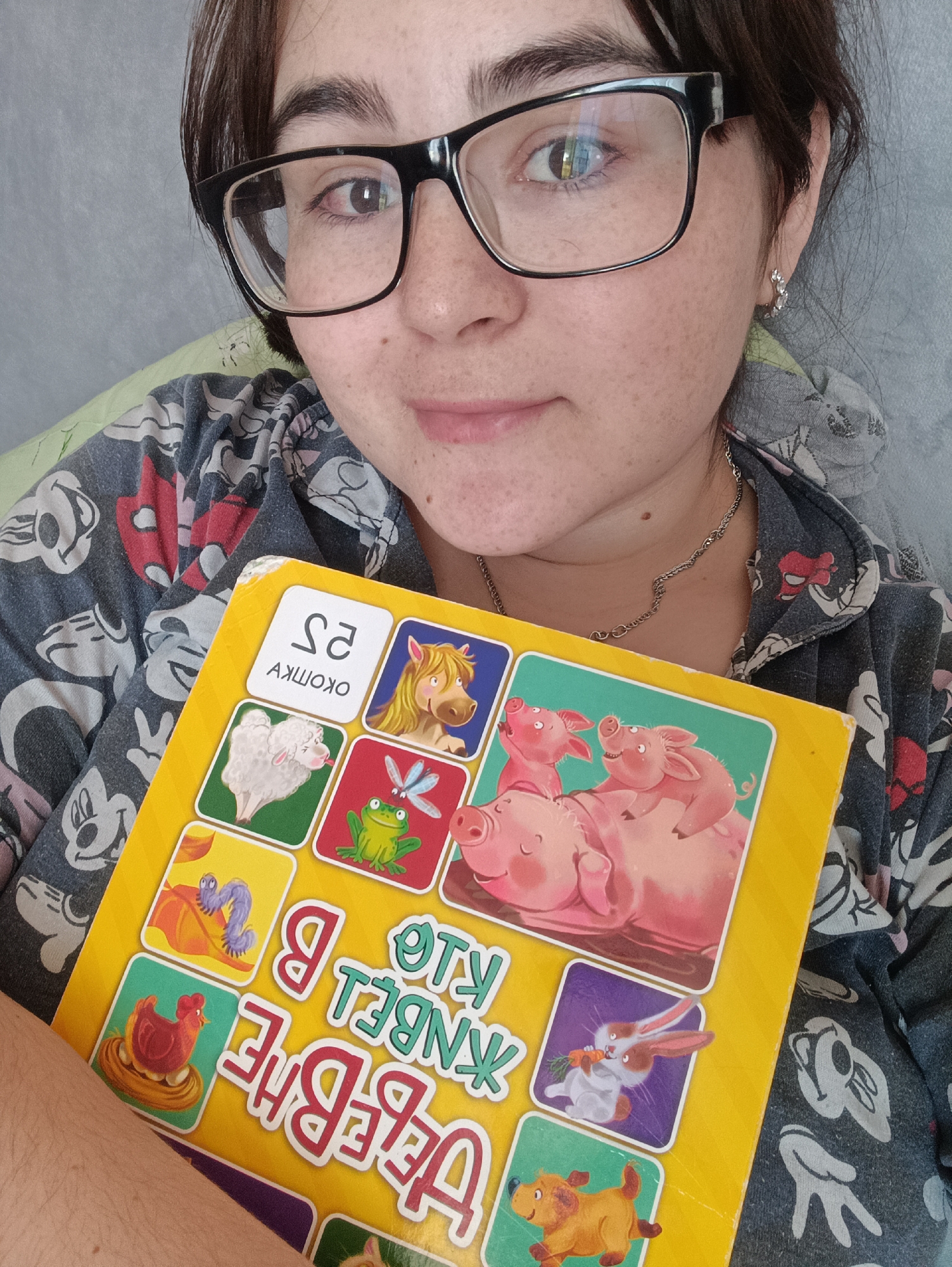 A fair-skinned person with dark hair and glasses is holding a yellow children's book with animal pictures. The person is wearing a patterned pajama top featuring Mickey Mouse and other characters. The text on the book is in a language other than English. The background is indistinct, but appears to be an indoor setting. The person's face is slightly flushed. No city, including Noviy, Russia is shown in the image.