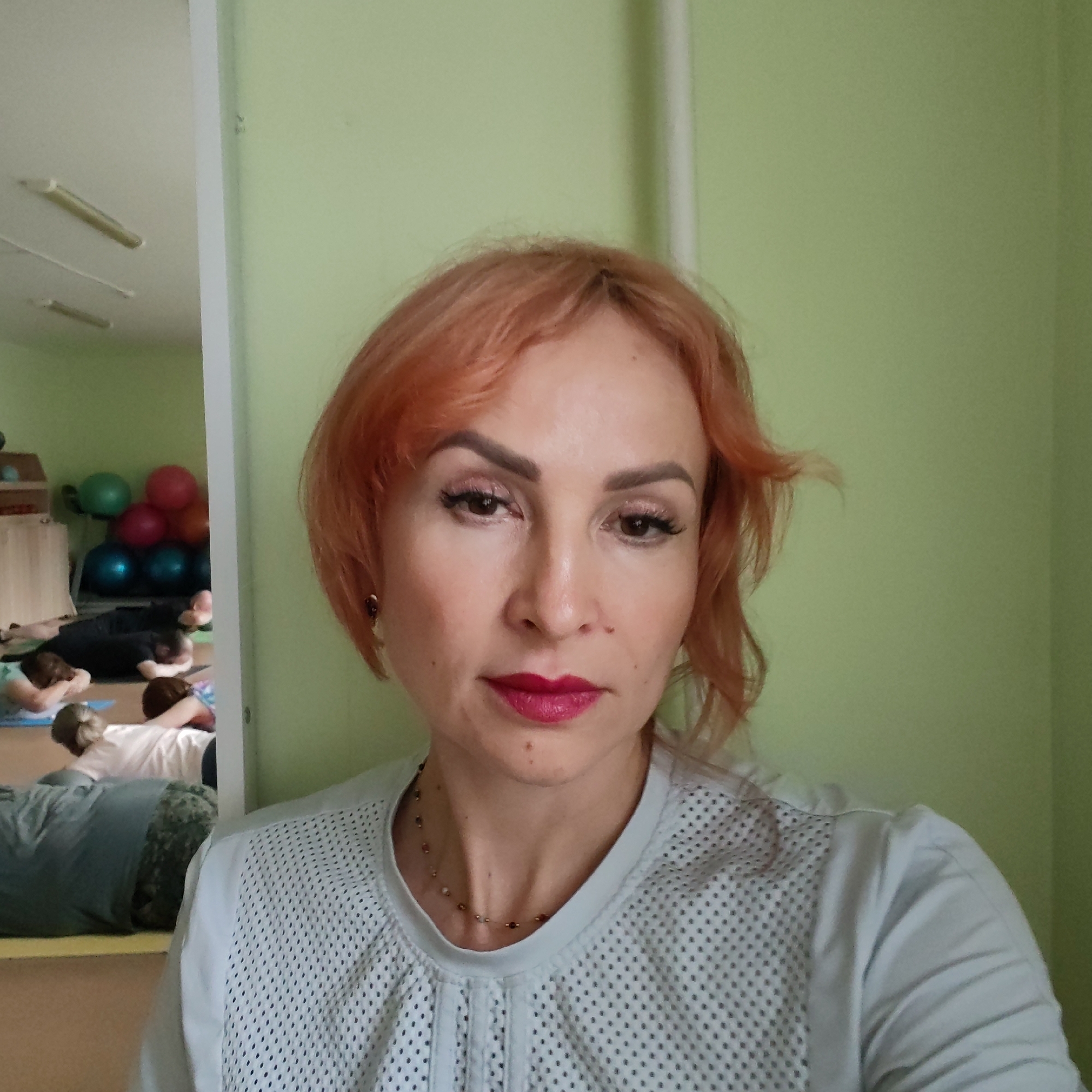 The image is a selfie of a woman with short, wavy, reddish-orange hair, light skin, and dark eyes. She is wearing prominent makeup, including dark eyeliner, defined eyebrows, and bright fuchsia lipstick. She has small earrings and a delicate, beaded necklace. Her top is light-colored, possibly white or light grey, with a textured or perforated front panel. Her expression is neutral to slightly serious.
Directly behind the woman is a plain, light green wall. To her left, a large mirror reflects another section of the room. In the reflection, several people are seen lying on yellow and blue exercise mats, their backs mostly facing the camera, indicating they are participating in a group fitness activity such as yoga or Pilates. Their faces are not visible. The reflected room also features light green walls, and stacked exercise balls in red, green, and blue are visible, along with what appears to be a wooden storage unit. Fluorescent light fixtures are visible on the ceiling in the reflection.
The scene depicts an indoor environment, likely a fitness or wellness studio in Chaikovsky, Russia, where a group exercise class is in progress. The time of day appears to be daytime, judging by the ambient lighting. No text is visible in the image.