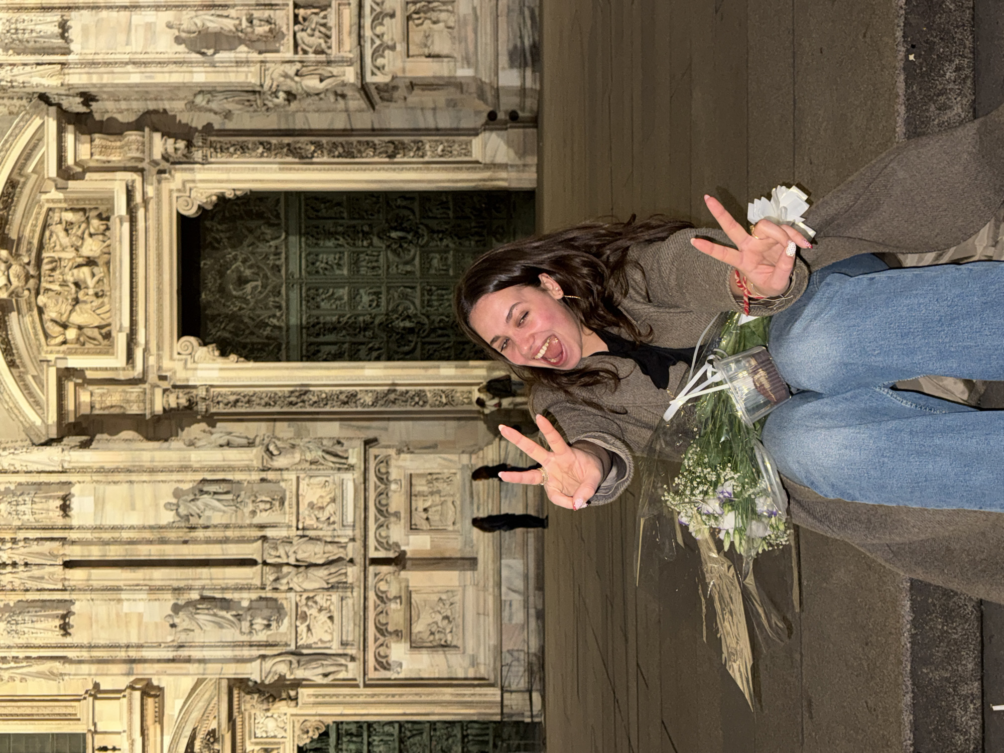 The image displays a young adult female seated on stone steps directly in front of the Duomo di Milano (Milan Cathedral) in Milan, Italy, at night. The woman has long, dark brown hair and is wearing a dark brown, textured long coat over blue jeans. She exhibits a wide, open-mouthed smile, with her eyes partially squinted, and both hands are raised, each forming a peace sign (V-sign). Her right hand displays a ring on the ring finger and light-colored nail polish with red polka dots on the ring finger. Resting on her lap are a bouquet of white and light purple flowers, wrapped in clear plastic, and a clear plastic cup containing a dark, possibly chocolate-based, item. The background features the elaborately carved and illuminated Gothic facade of the cathedral, including multiple statues in niches, ornate stonework, and a large bronze-colored main entrance door. Several indistinct figures are visible standing on the paved area in the mid-ground, further behind the main subject.