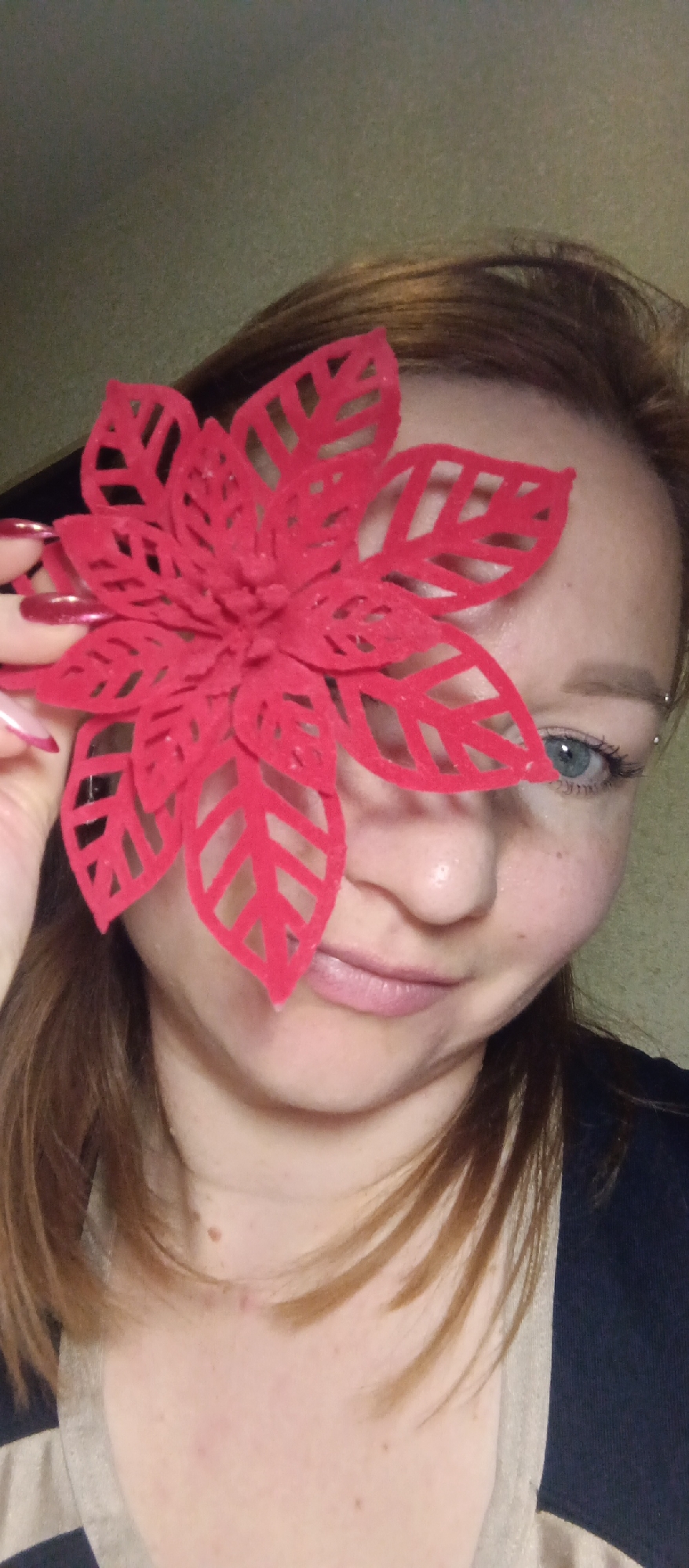 A close-up, eye-level indoor portrait features a woman holding a large, intricately cut red felt flower in front of her face, partially obscuring it.
The woman has light brown or auburn hair that frames her face. Her right eye, visible behind a petal, is blue and appears to be looking at the camera. Her complexion is light, with some freckles or moles visible on her cheek and neck. Her lips are a soft pink, slightly parted in a gentle smile. She is wearing a dark top, with a cream-colored, possibly striped, garment visible on her left side. Her right hand, which holds the flower, has long, pointed red nails, suggesting a manicure. Two small studs are visible near the outer corner of her right eyebrow.
The red felt flower is prominent, designed with multiple layers of petals featuring delicate cut-out leaf patterns, resembling a poinsettia. It covers her left eye and much of her nose and mouth.
The background is out of focus, appearing as a plain wall in a muted greenish-gray or brownish-gray tone. The lighting is soft, suggesting an indoor setting. There is no visible text in the image.