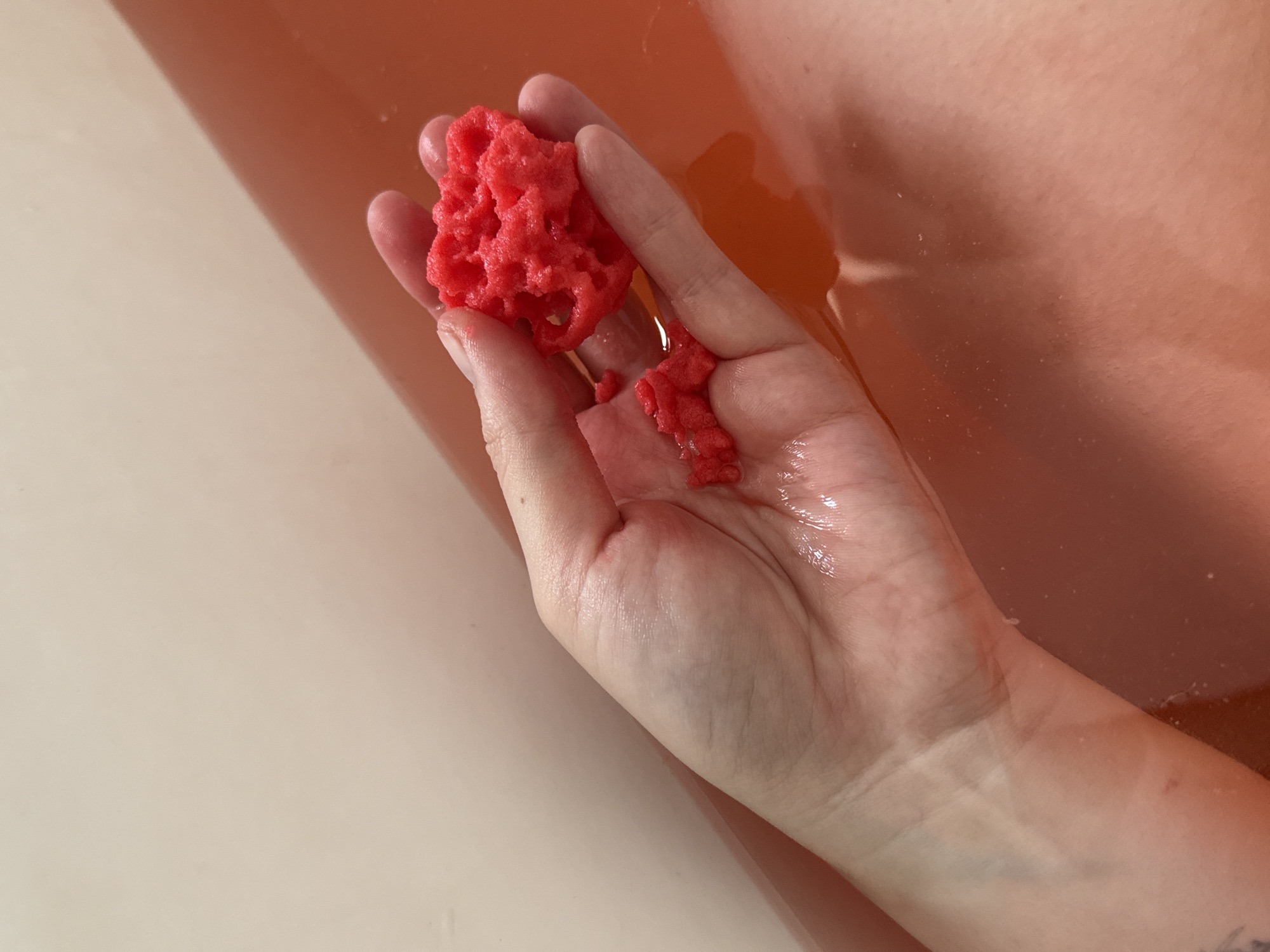 A close-up shot shows a person's hand holding a red sponge over a bathtub filled with reddish-brown water. The bathtub is white with a slight curve. Part of the person's body is also visible in the water. There is no indication of the location being Petah Tikva, Israel, or any identifiable location.