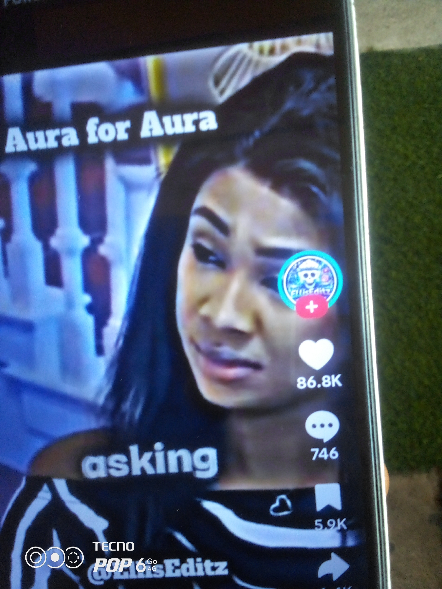 The image shows a close-up of a smartphone screen displaying a video. The video features a woman with dark hair and makeup, looking towards the camera. Text overlays on the screen read "Aura for Aura" at the top, and "asking" towards the bottom. On the right side of the screen, there are social media interaction icons, including a heart with "86.8K" below it, a speech bubble icon with "746" below it, and a bookmark icon with "5.9K" below it. A circular profile picture with a skull inside is also visible near the woman's face. The lower part of the phone screen shows "TECNO POP 6 Go 4G" and "@EllasEditz". The background of the image shows the phone held in front of a green, textured surface, possibly artificial turf. There is no visible indication of the city of Howayi, Nigeria within the image content.