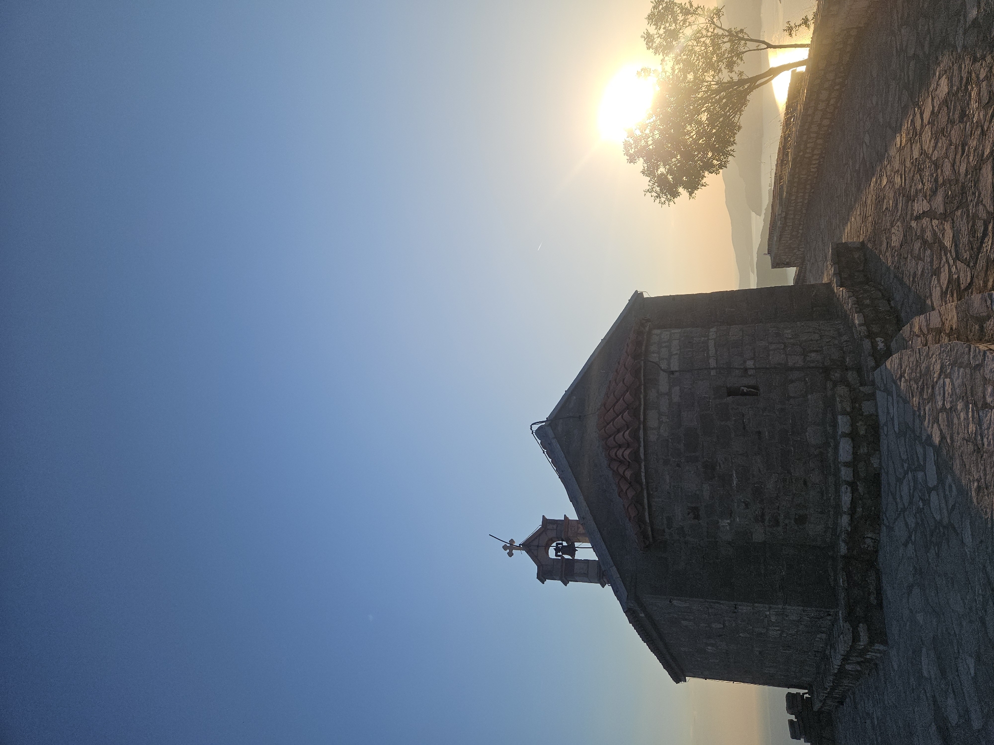 A stone chapel with a red-tiled roof stands on a paved stone pathway overlooking the sea. The chapel has a bell tower topped with a golden cross. A tree with sparse leaves is silhouetted against the bright, setting sun on the right side of the image. The sun casts long shadows across the pathway. The sea and distant hills are visible in the background. The scene appears to be a viewpoint on Sveti Stefan, Montenegro. There are no people visible in the image.