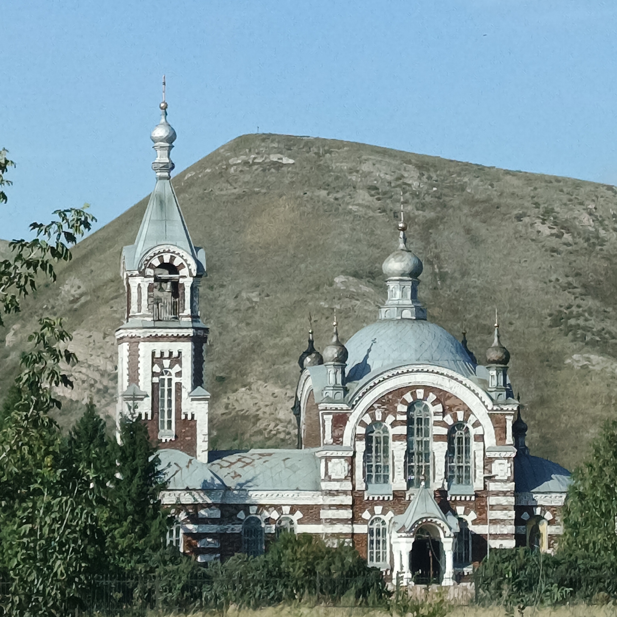 The image shows a red brick church with white decorative elements and a large, light-blue domed roof. A bell tower with a similar roof structure stands to the left of the main building. The church is surrounded by green trees in the foreground and the middle ground. Behind the church and trees, a gently sloping, barren hill occupies the background, with a clear blue sky above. The scene is brightly lit, suggesting daylight, and the overall impression is one of a historic religious structure in a natural, possibly rural or semi-rural, setting. The specific location in Андреевка, Russia, is not visually discernible from the image itself but is provided as context. There are no people or specific actions depicted.